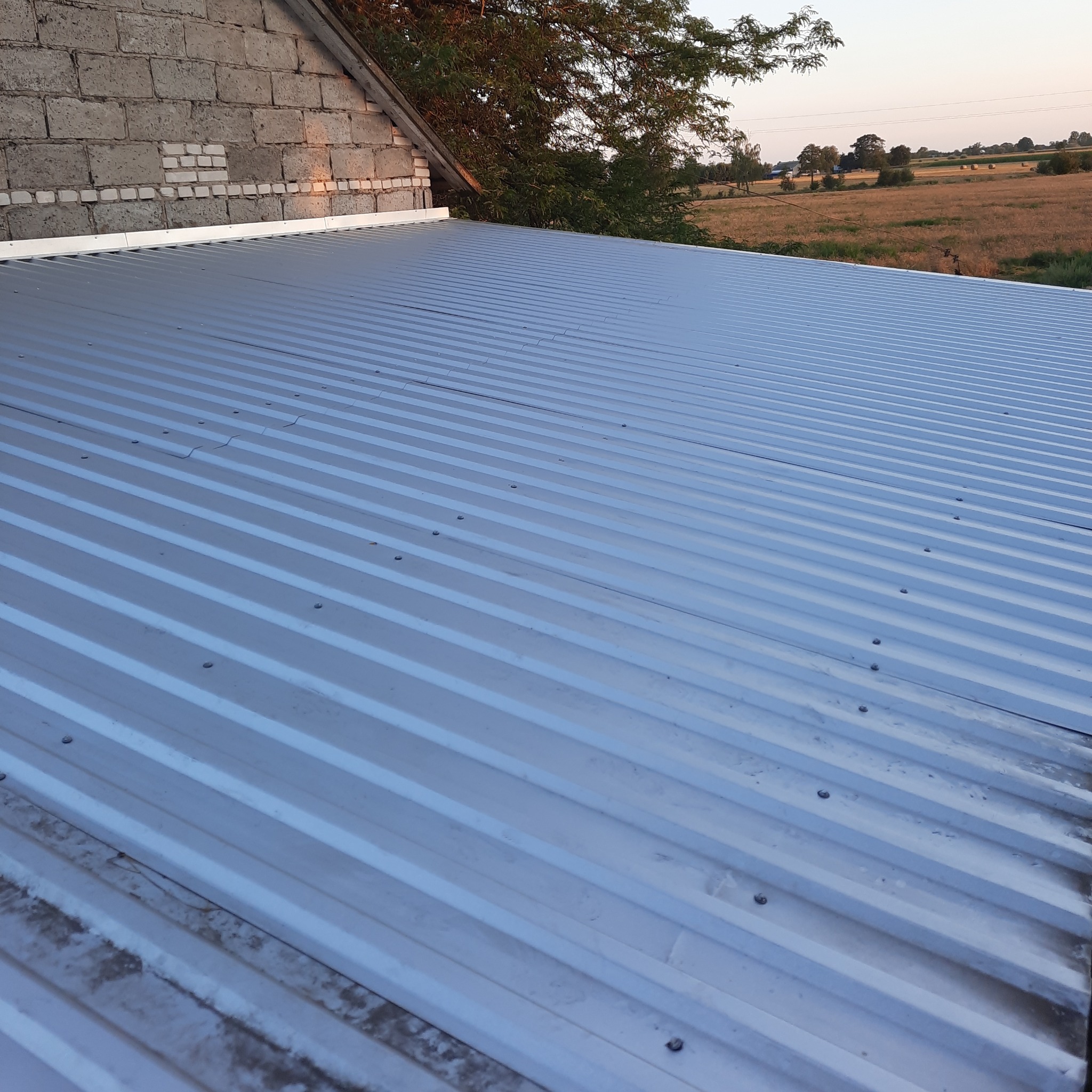 Nowy, srebrny dach z blachy trapezowej, zamocowany na budynku z ceglaną ścianą szczytową i widokiem na pola uprawne w oddali.