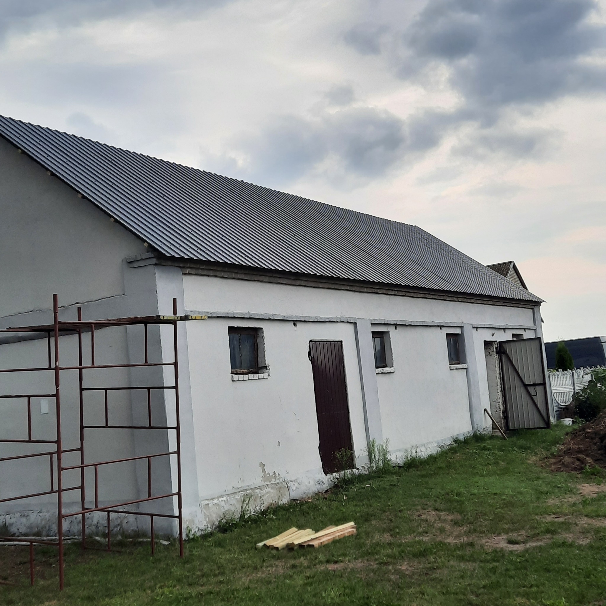 Budynek gospodarczy z nowym, szarym dachem z blachy falistej, widoczne rusztowanie i ułożone deski na trawie.