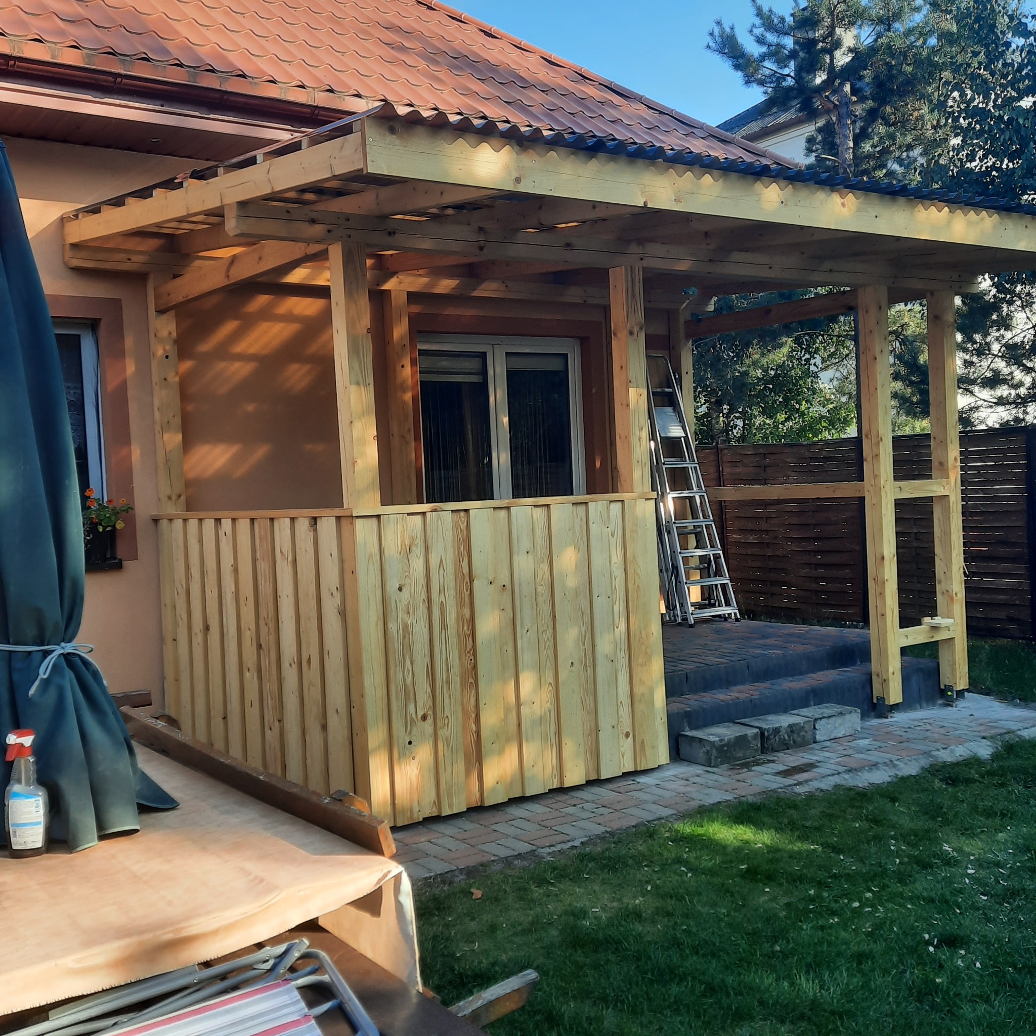 Budowa drewnianej werandy z częściowo zamontowaną boazerią, widoczna drabina oparta o konstrukcję i fragment istniejącego dachu z czerwonej dachówki.