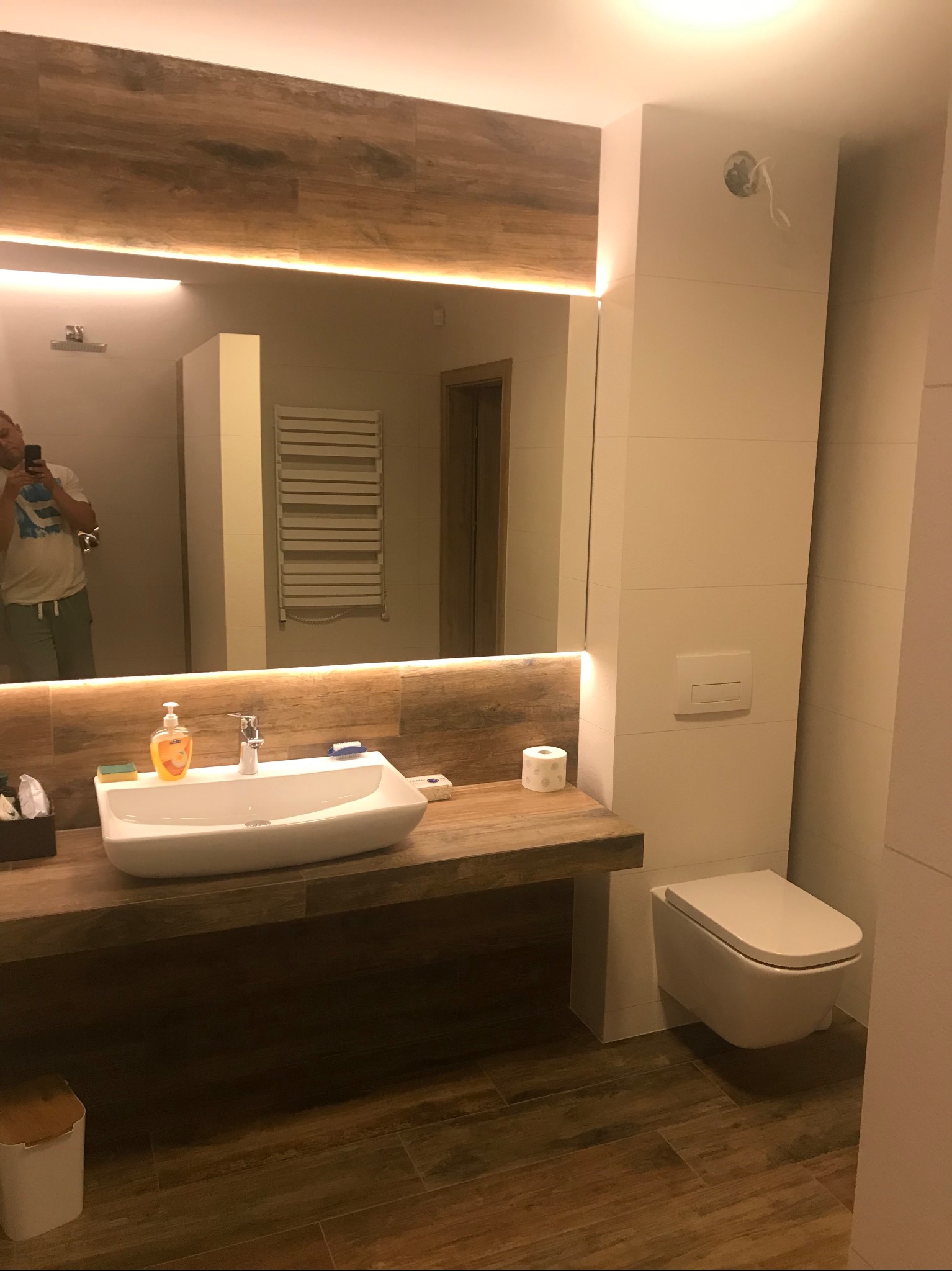 Nowoczesna łazienka z drewnopodobnymi płytkami na podłodze i ścianie, umywalka nablatowa na drewnianym blacie z oświetleniem LED pod lustrem, wisząca toaleta, widoczne akcesoria łazienkowe...