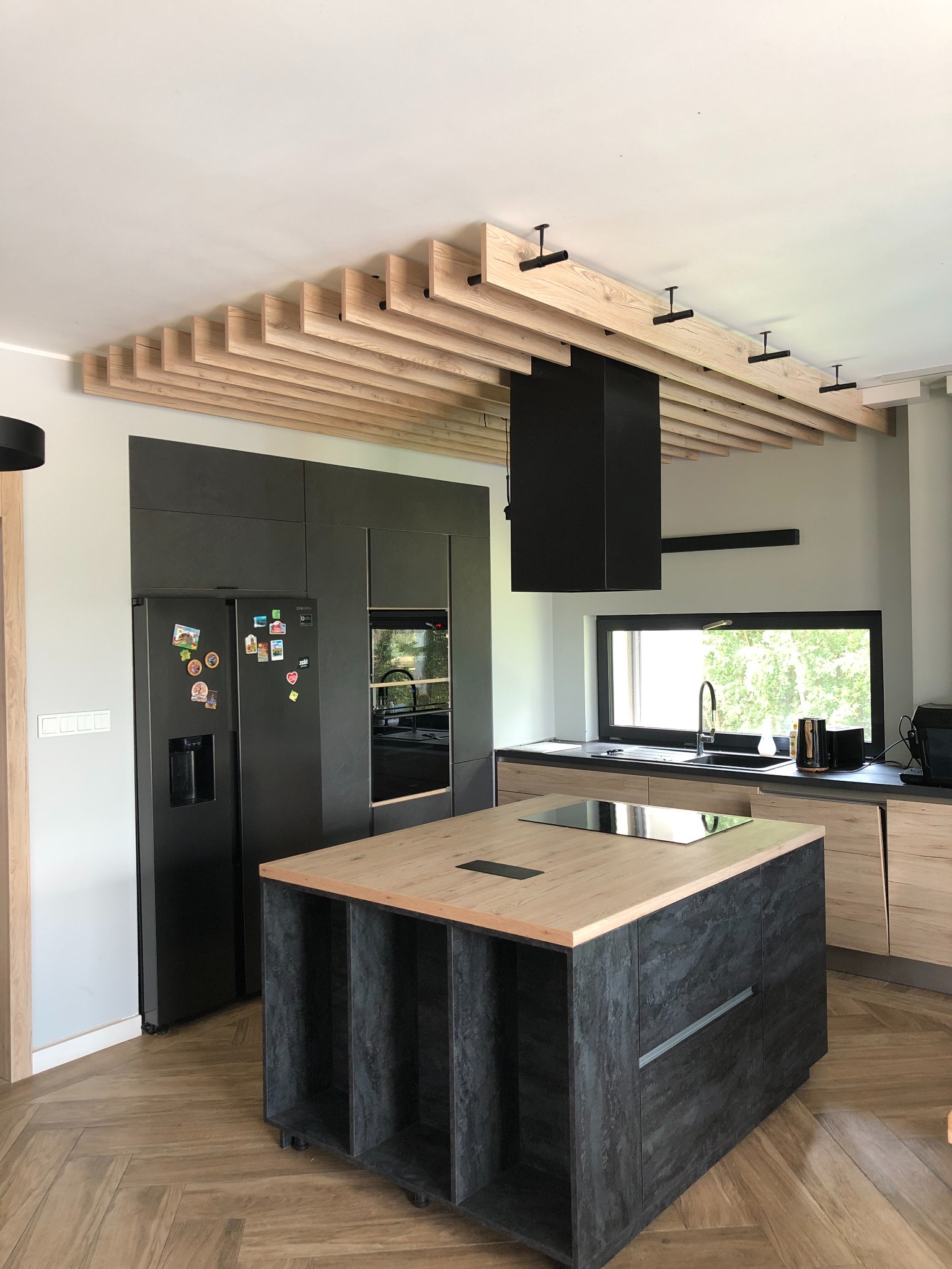 Nowoczesna kuchnia z wyspą i drewnianym sufitem lamelowym, czarne AGD, parkiet w jodełkę.