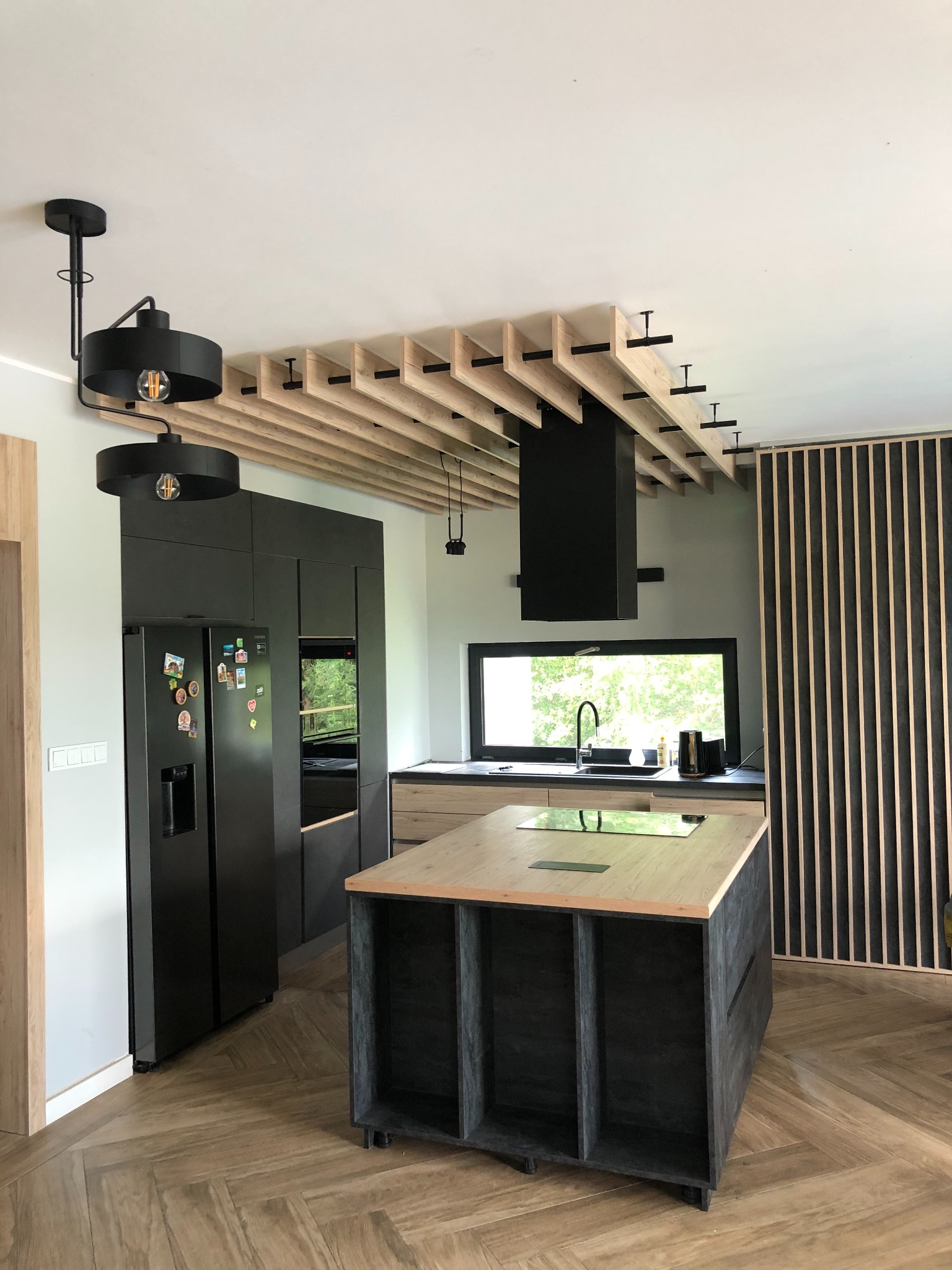Nowoczesna kuchnia z czarnymi szafkami, wyspą z blatem w kolorze drewna i dekoracyjnym sufitem z drewnianych listew z czarnymi mocowaniami, oświetlenie w stylu industrialnym.