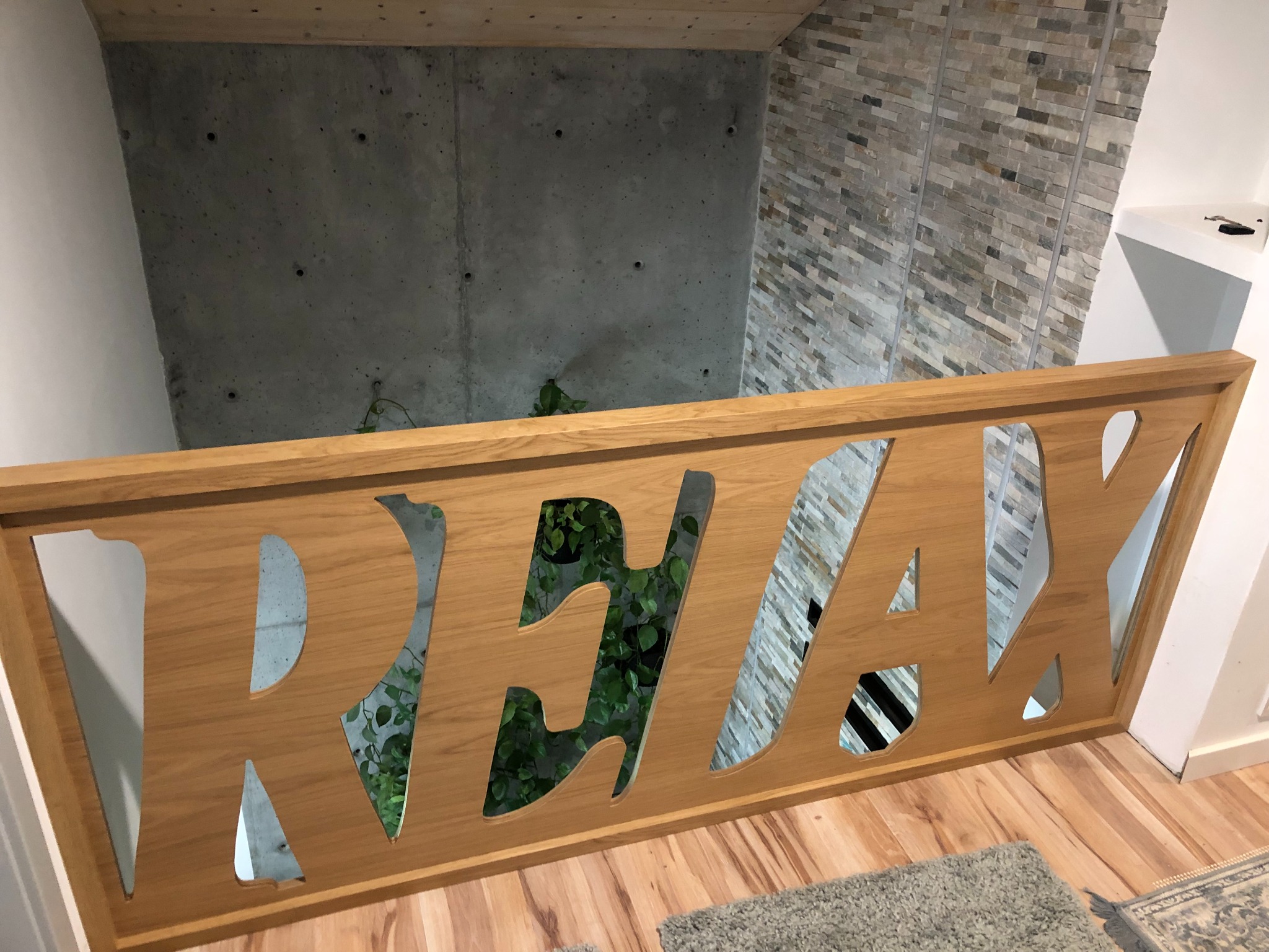 Drewniana balustrada schodów z wyciętym napisem 'RELAX', widok z góry na betonową ścianę i ścianę z kamienia dekoracyjnego, widoczna roślina doniczkowa.