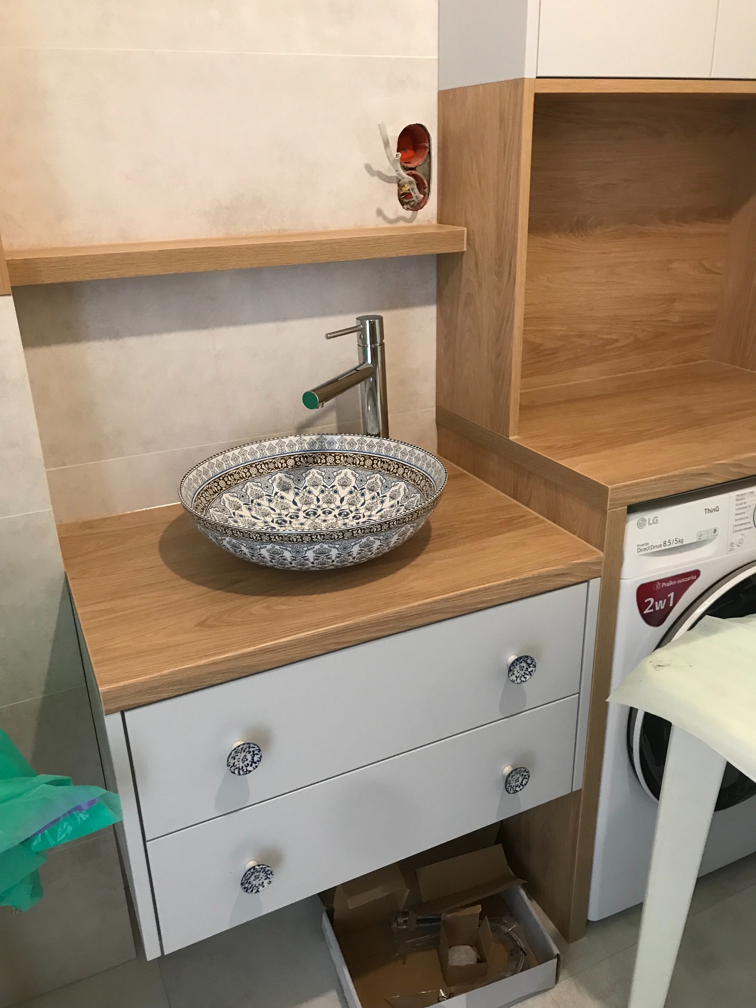 Umywalka nablatowa w etniczne wzory na szafce z dwiema szufladami i ozdobnymi gałkami, obok pralko-suszarka i otwarte półki z drewnianym wykończeniem.