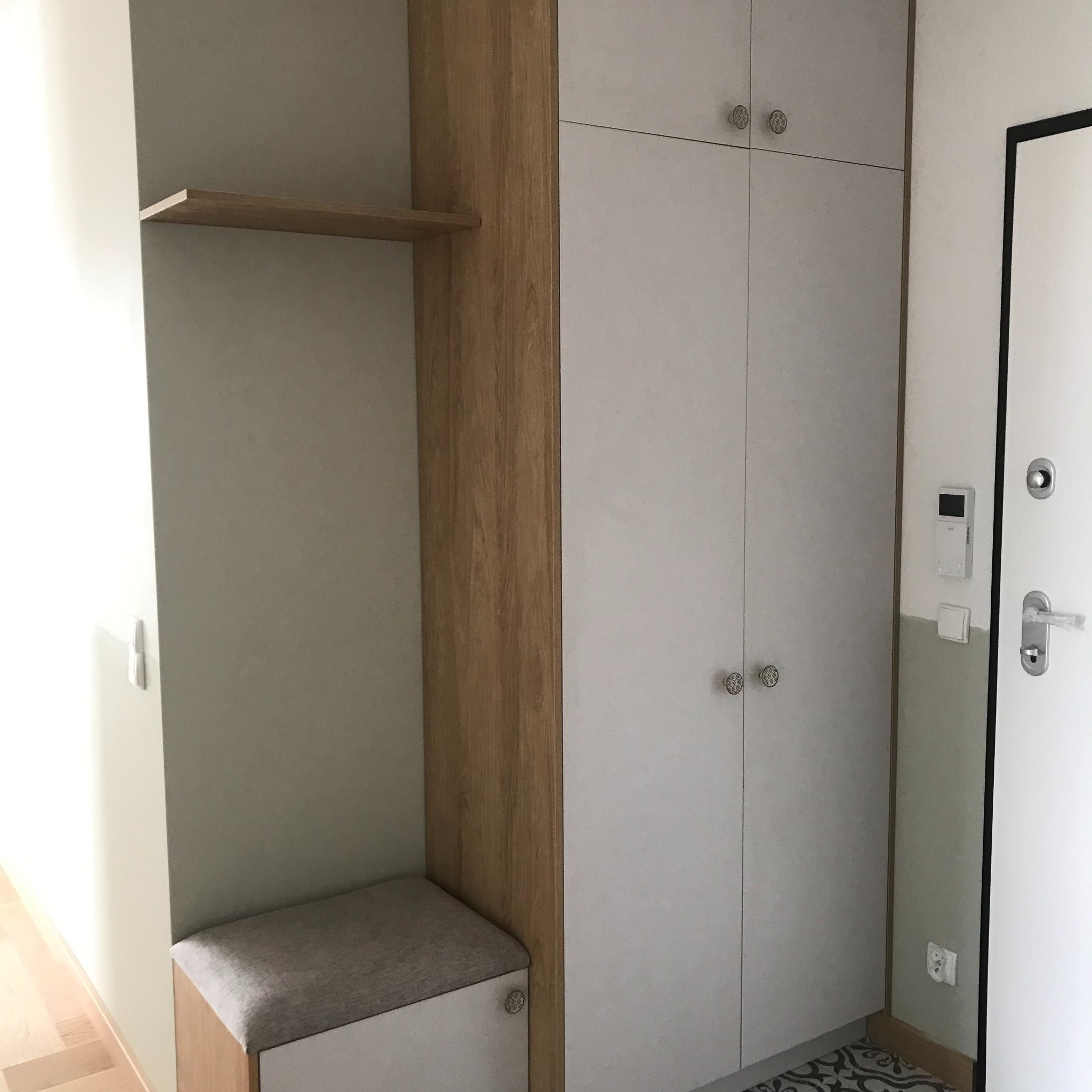 Jasnoszara szafa z drewnianymi akcentami i siedziskiem w przedpokoju, widoczne dekoracyjne gałki i wzorzysta podłoga, przy drzwiach wejściowych.