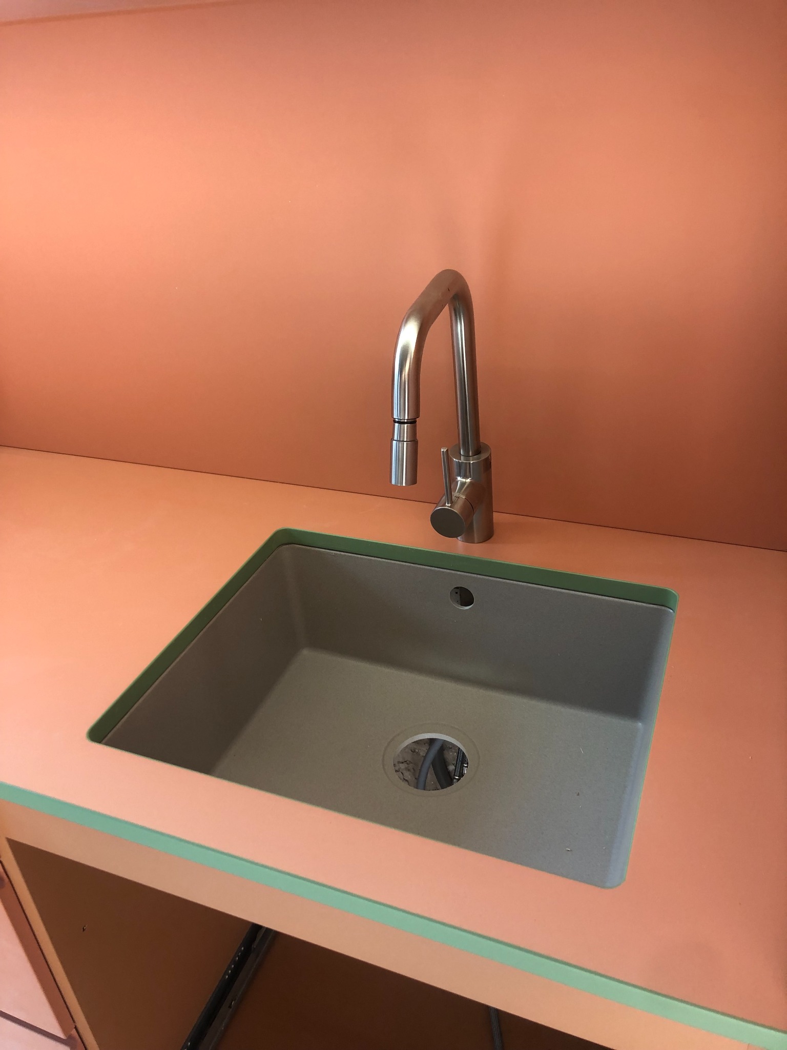 Nowoczesny zlewozmywak wpuszczany w blat o brzoskwiniowym odcieniu, z chromowaną baterią. Widoczne podłączenia hydrauliczne pod spodem.