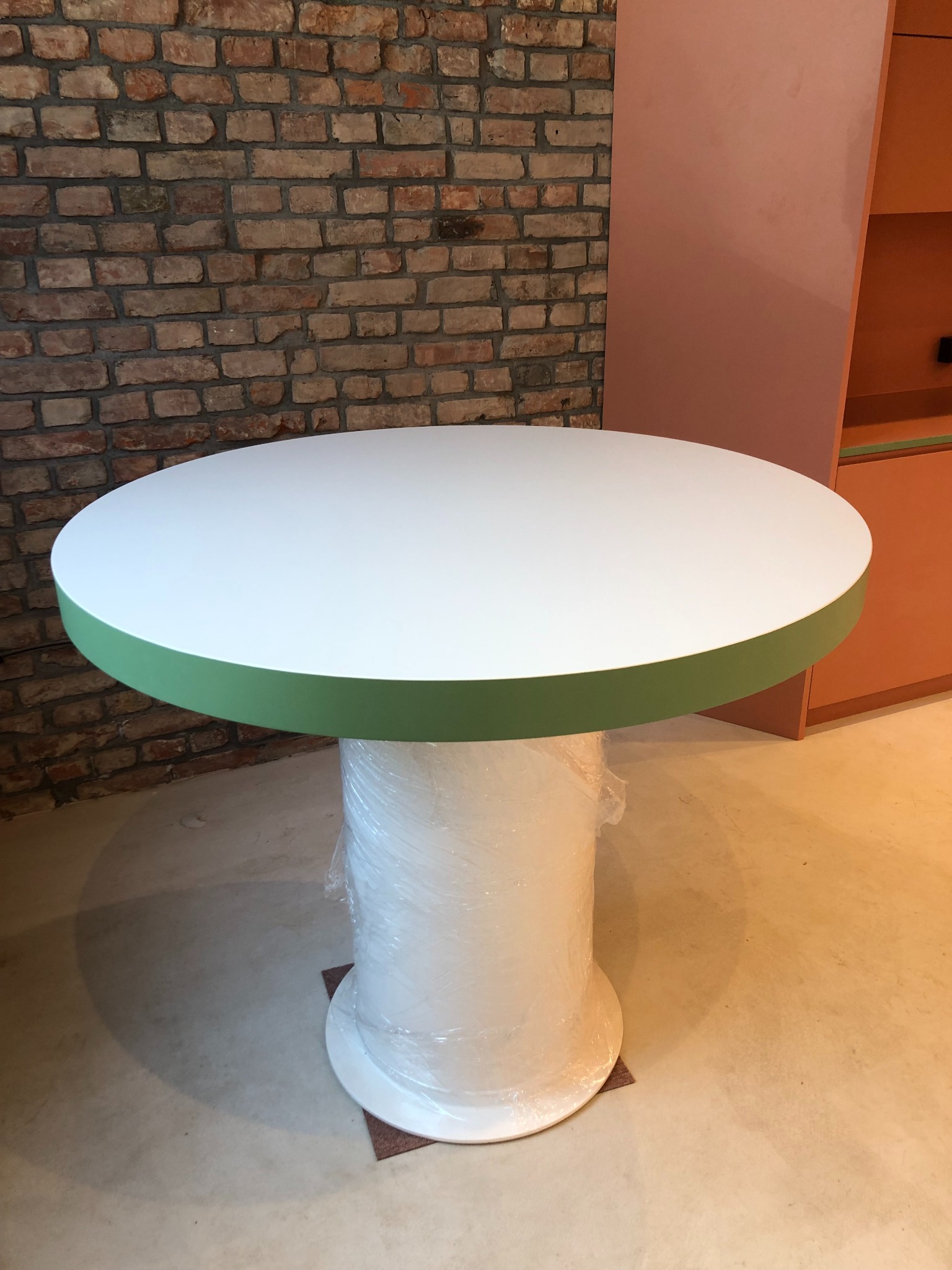 Okrągły stół z białym blatem i zielonym obrzeżem, kolumna owinięta folią ochronną, na tle ceglanej ściany i pomarańczowych mebli.