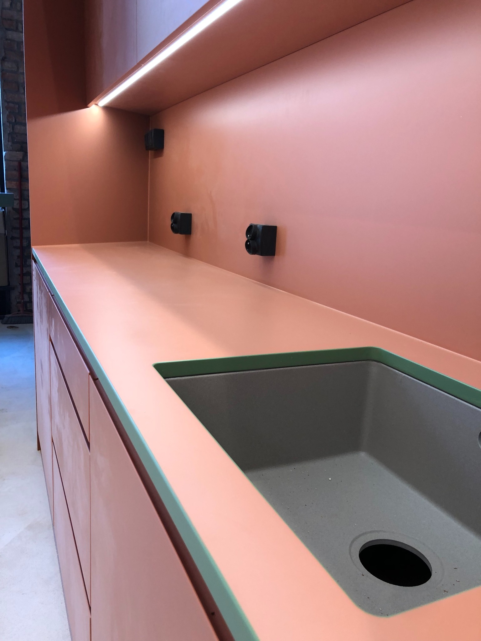 Nowoczesna kuchnia w Gdańsku z szafkami w kolorze brzoskwiniowym, szarym zlewem i zielonymi obramowaniami, oświetleniem LED pod szafkami wiszącymi i gniazdkami elektrycznymi na ścianie.