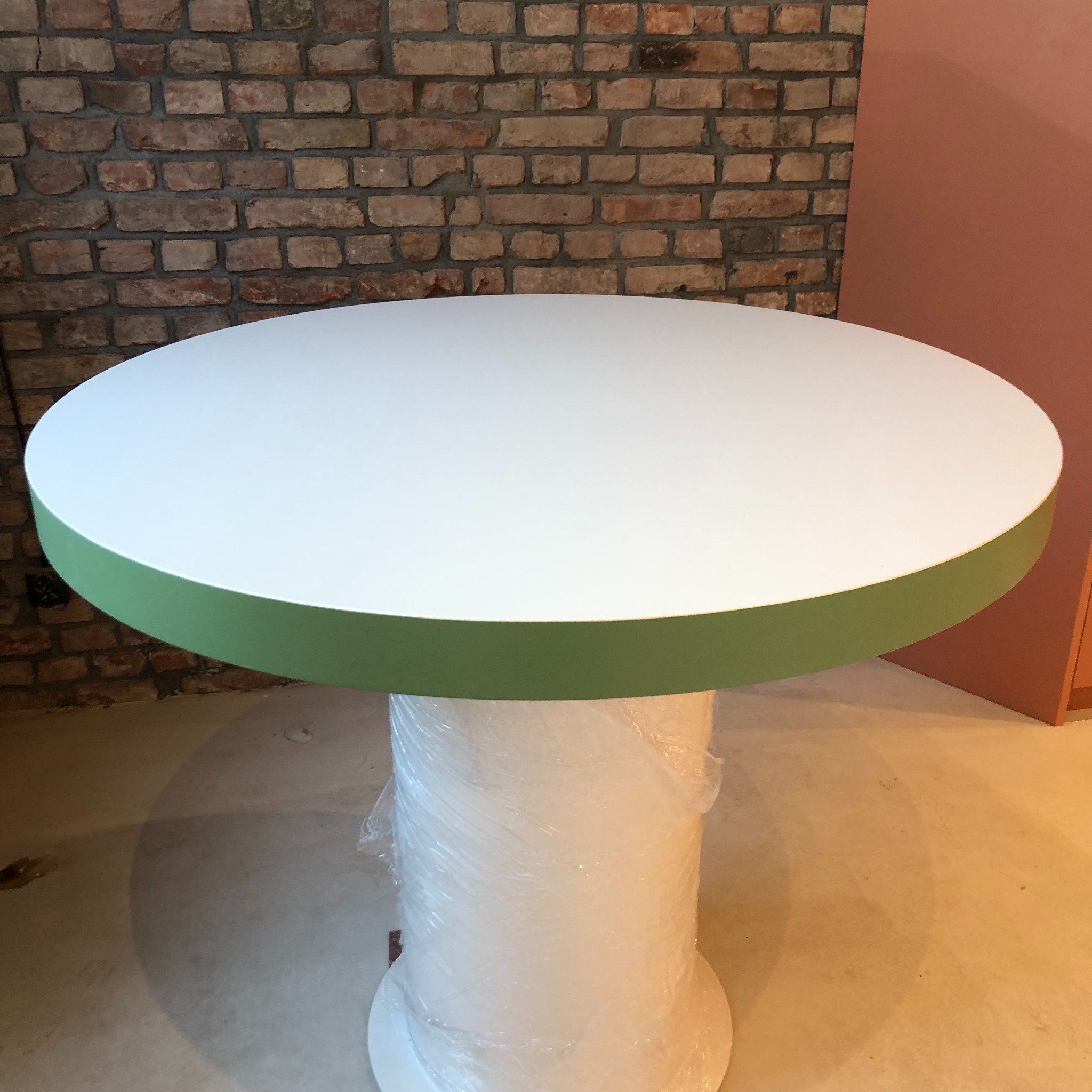 Okrągły blat stołu z białym wykończeniem i zielonym obrzeżem, kolumna owinięta folią ochronną, tło z ceglanej ściany i pomarańczowego panelu.