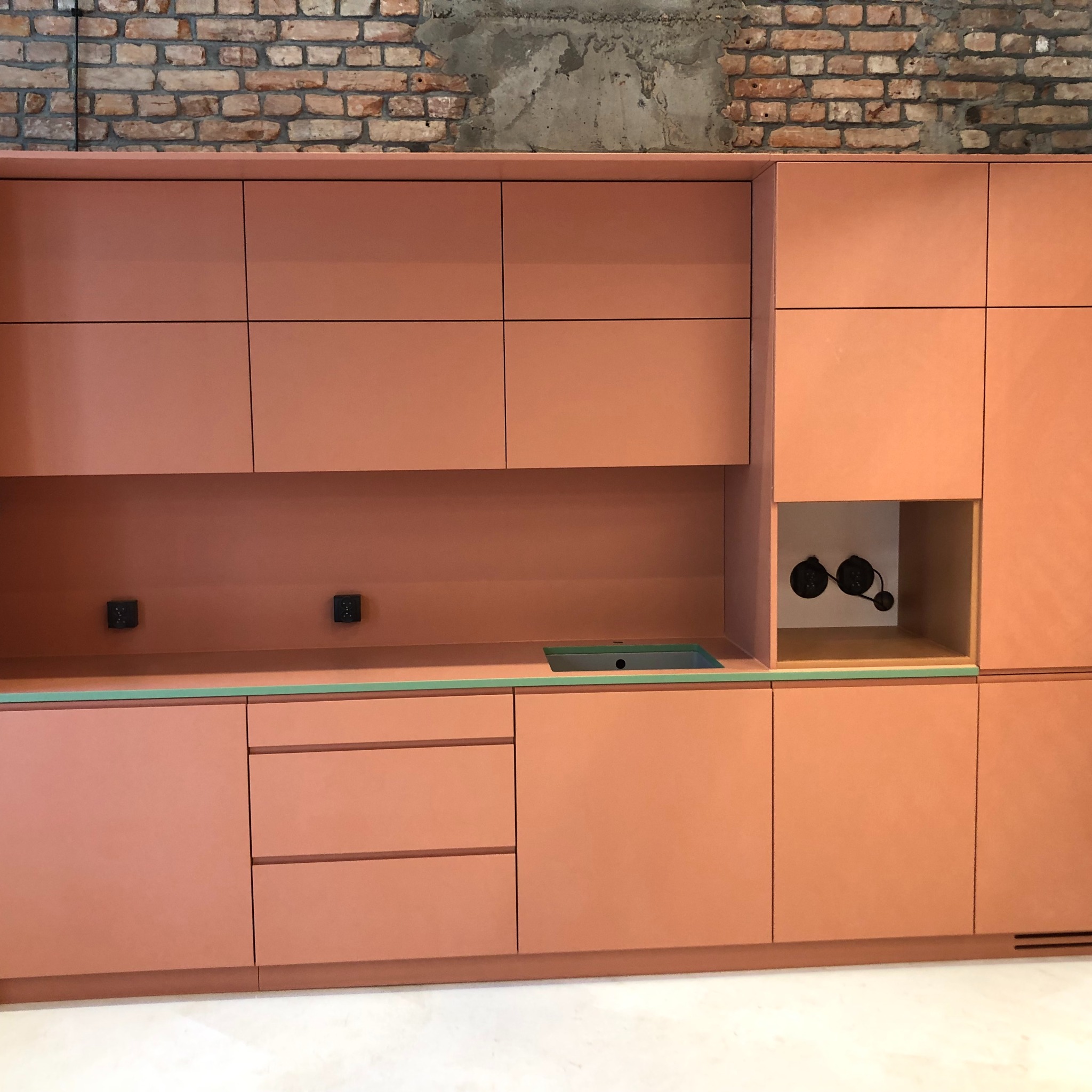 Nowoczesna zabudowa kuchenna w odcieniach terakoty z minimalistycznym zlewem i widocznymi gniazdkami elektrycznymi na tle surowej ściany z cegły i betonu.