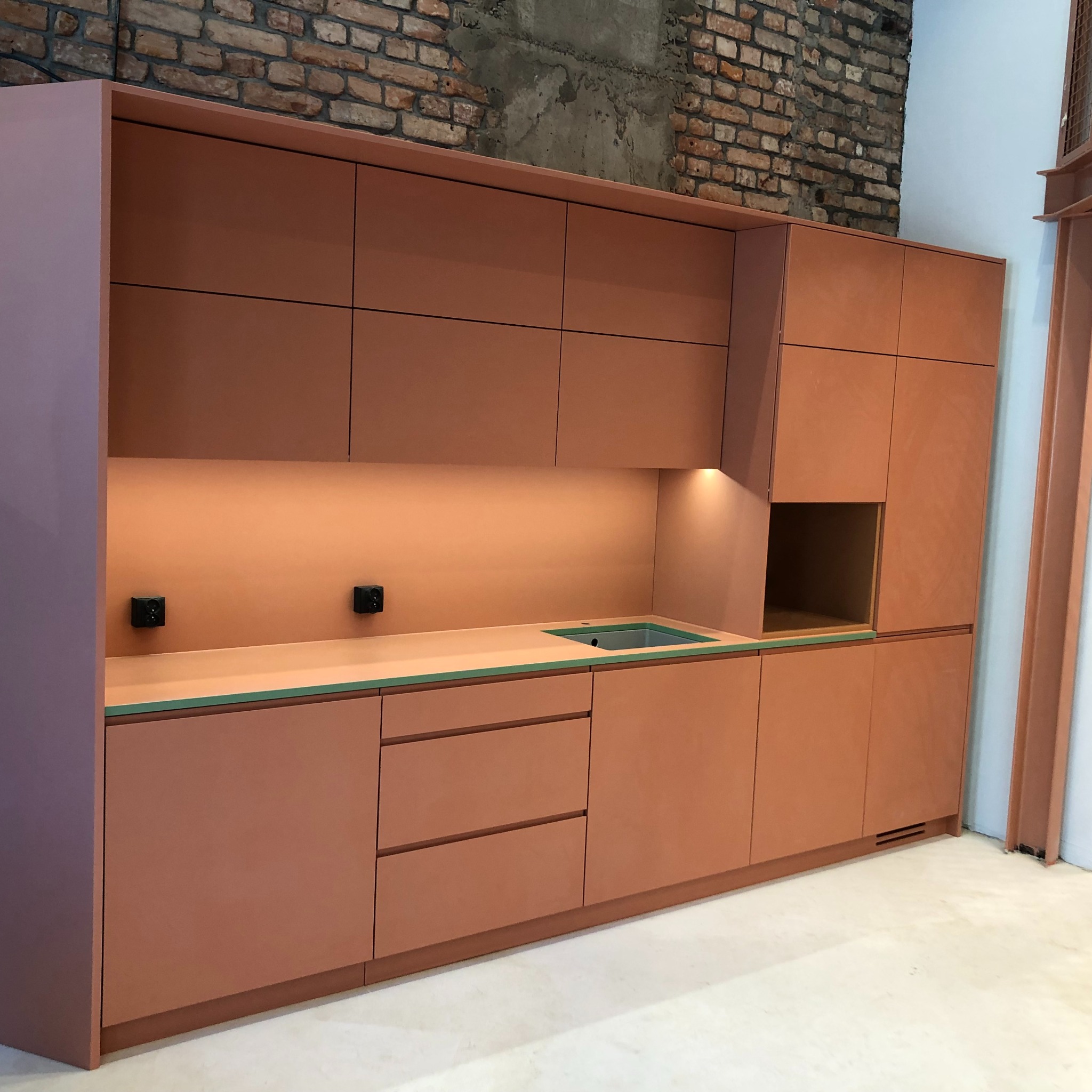 Nowoczesna zabudowa kuchenna w odcieniach terakoty z minimalistycznym blatem w kolorze zielonym, zlewem i oświetleniem punktowym, na tle surowej ściany z cegły.