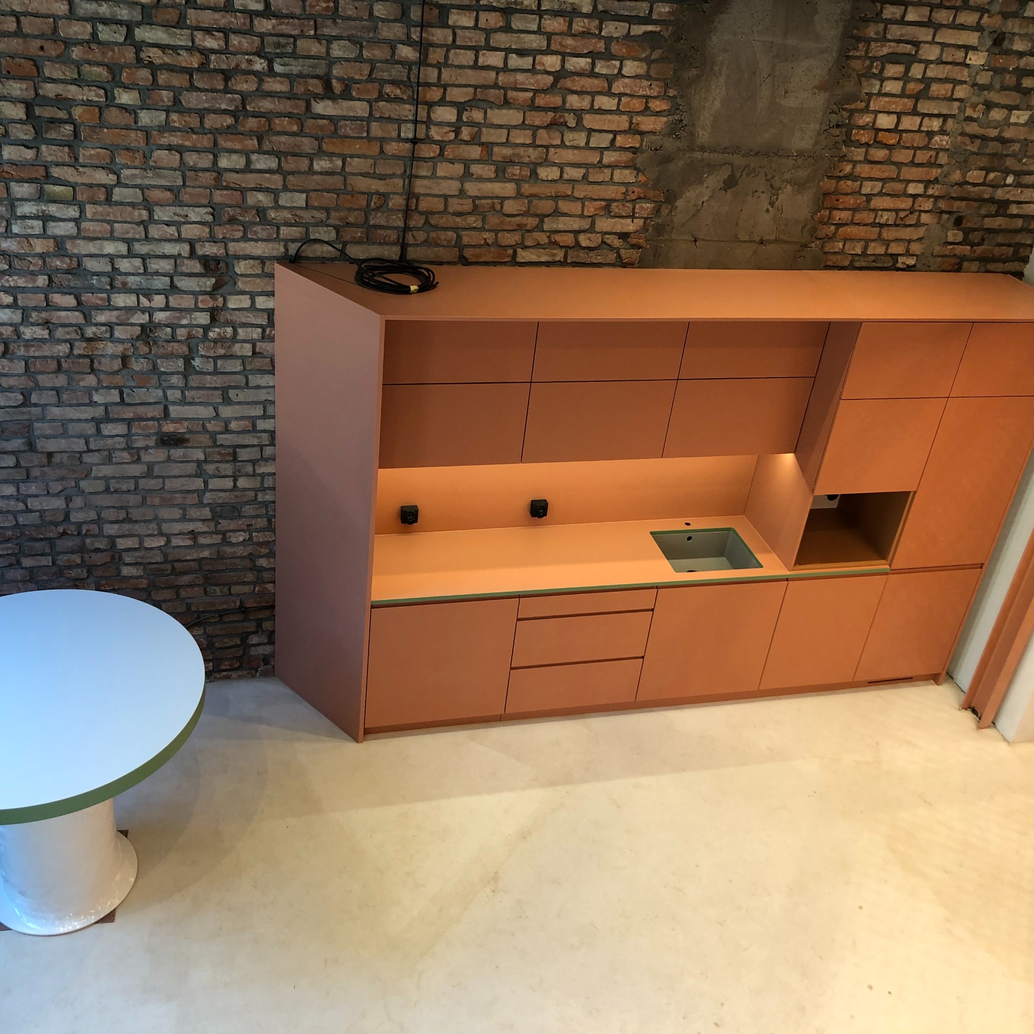 Nowoczesna zabudowa kuchenna w odcieniach terakoty z wbudowanym oświetleniem, umieszczona na tle ściany z czerwonej cegły, obok okrągły stół z białym blatem.