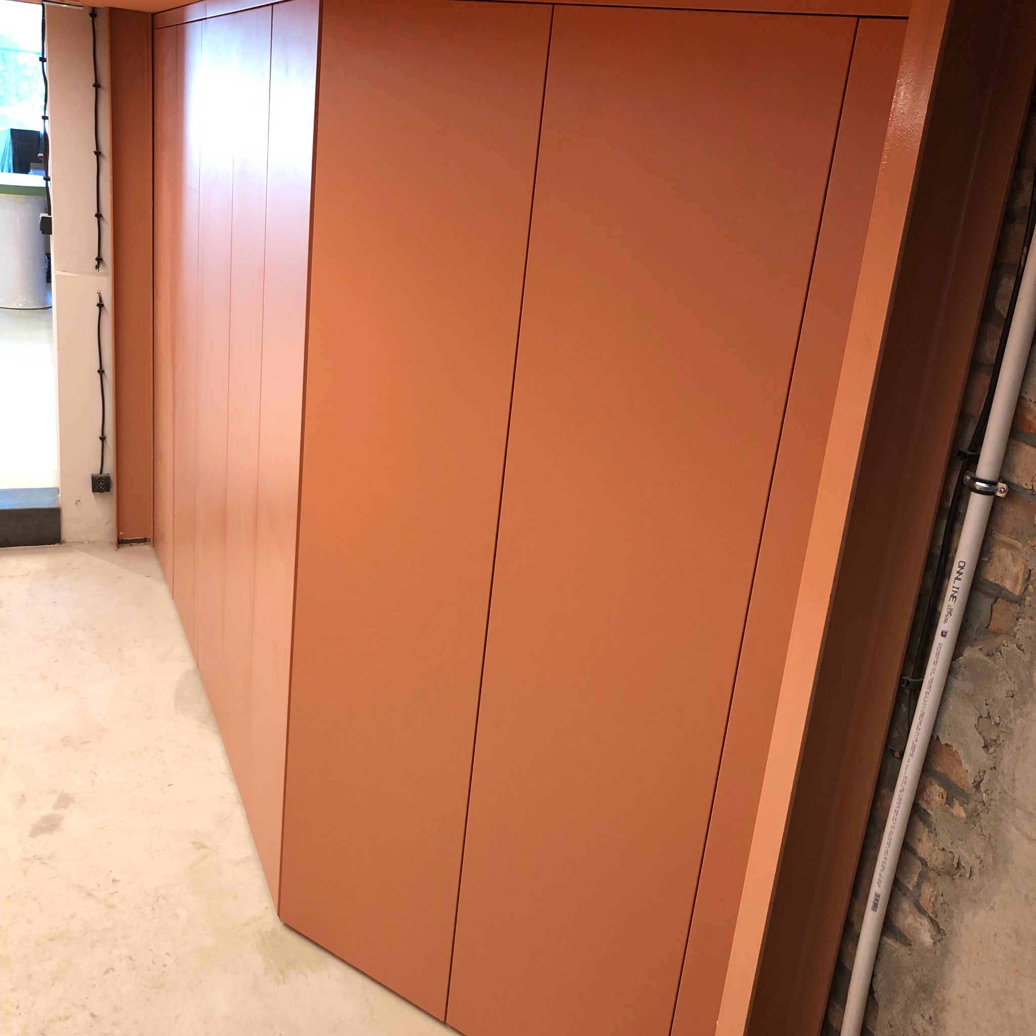 Pomarańczowa zabudowa meblowa wzdłuż ściany, widoczne pionowe podziały paneli, betonowa podłoga i fragment instalacji elektrycznej na ścianie.