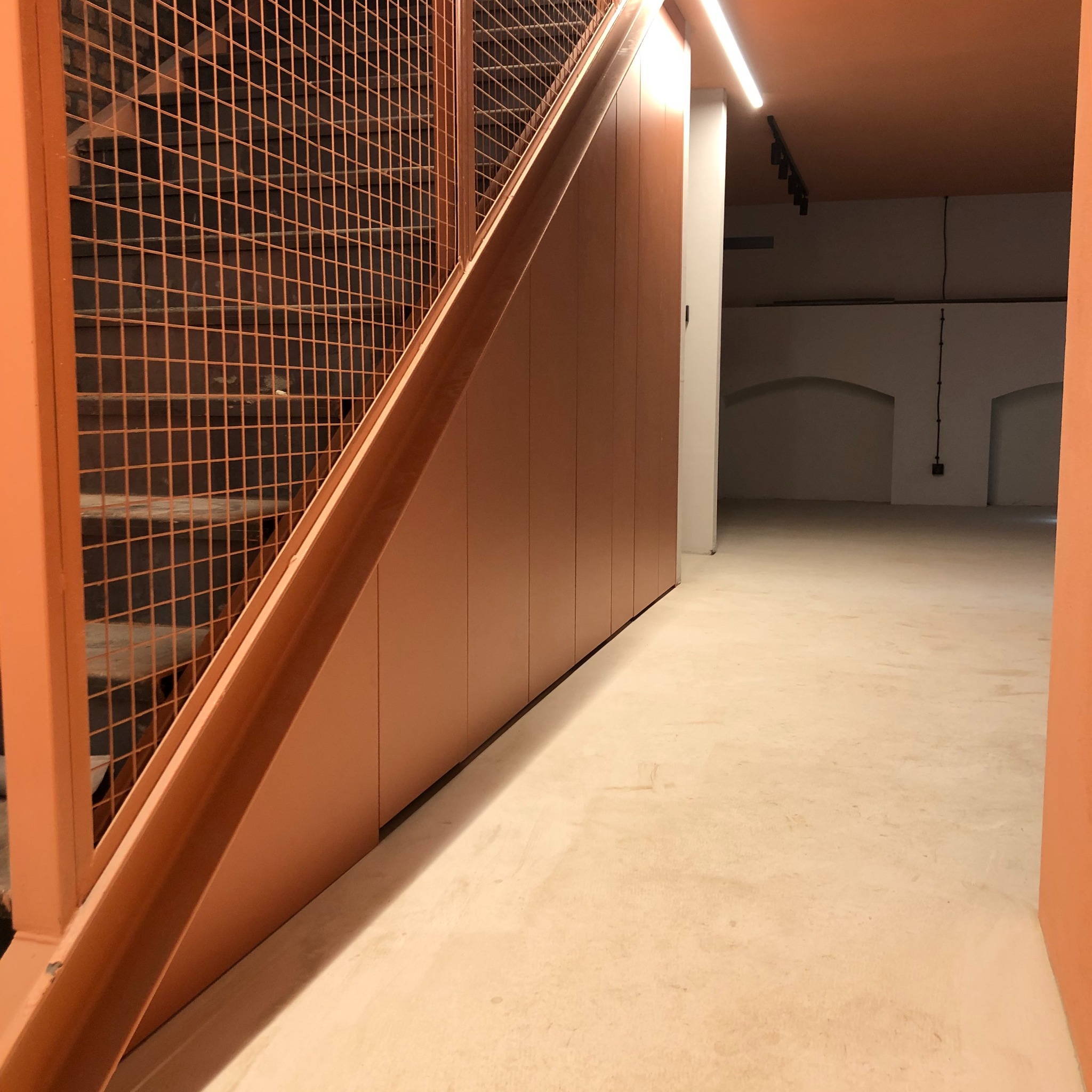Pomarańczowe szafy wnękowe pod schodami z metalową kratą, widok z dołu na puste pomieszczenie z łukowatymi wnękami w tle.