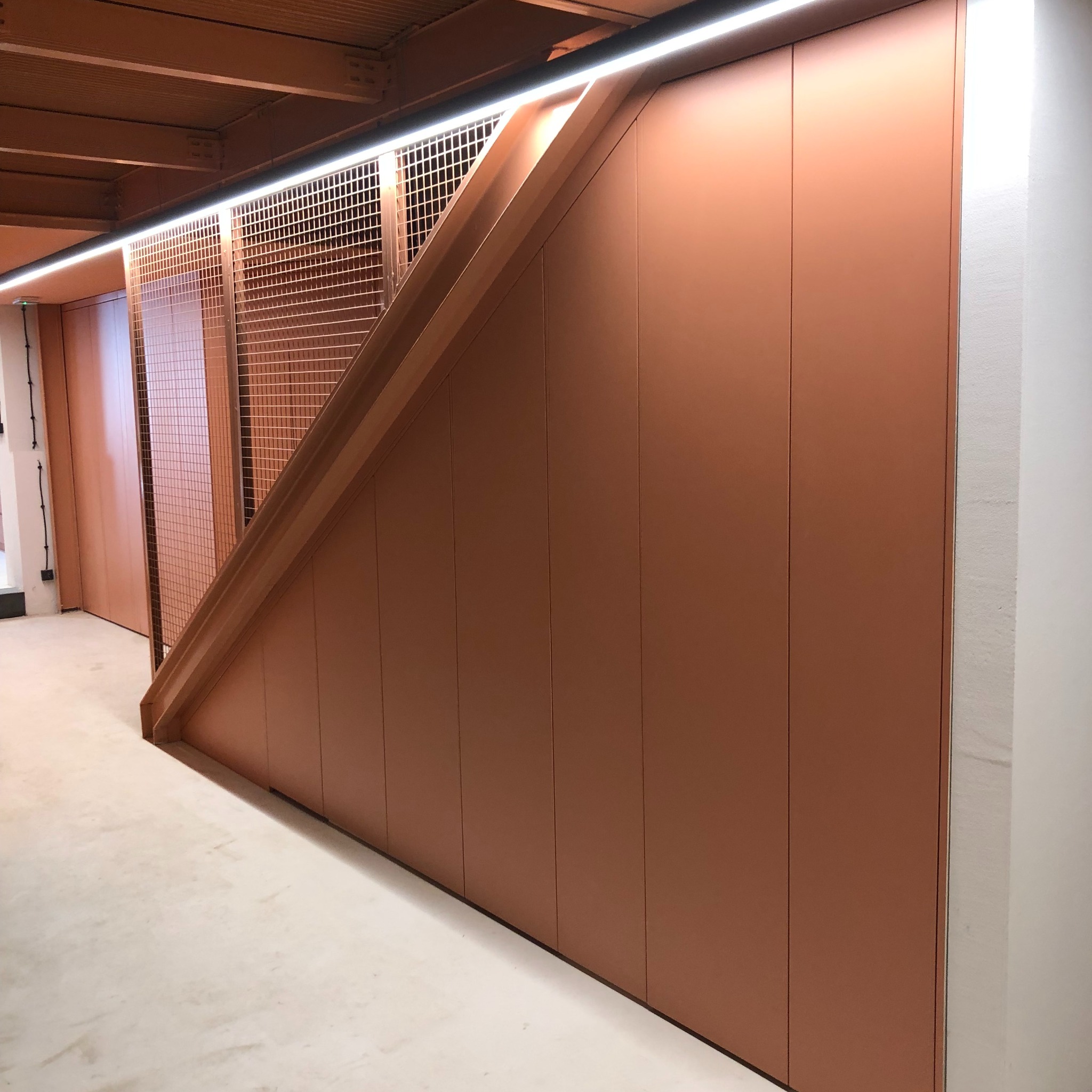 Nowoczesna szafa wnękowa w kolorze terakoty, zintegrowana ze schodami i metalową siatką zabezpieczającą, oświetlona liniowym światłem LED.