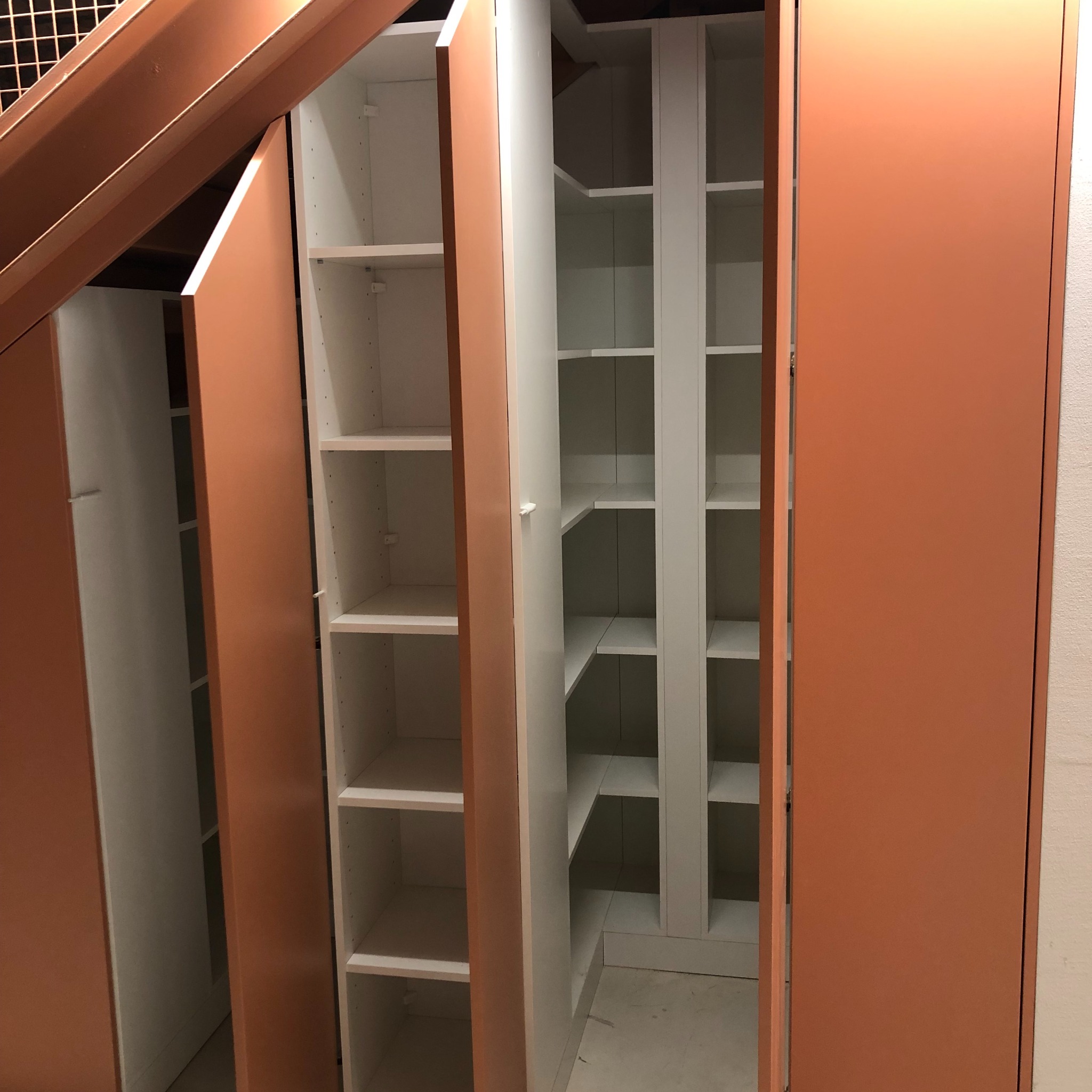 Otwarta szafa wnękowa z białymi półkami i pomarańczowymi drzwiami, widoczna część konstrukcji pod schodami.