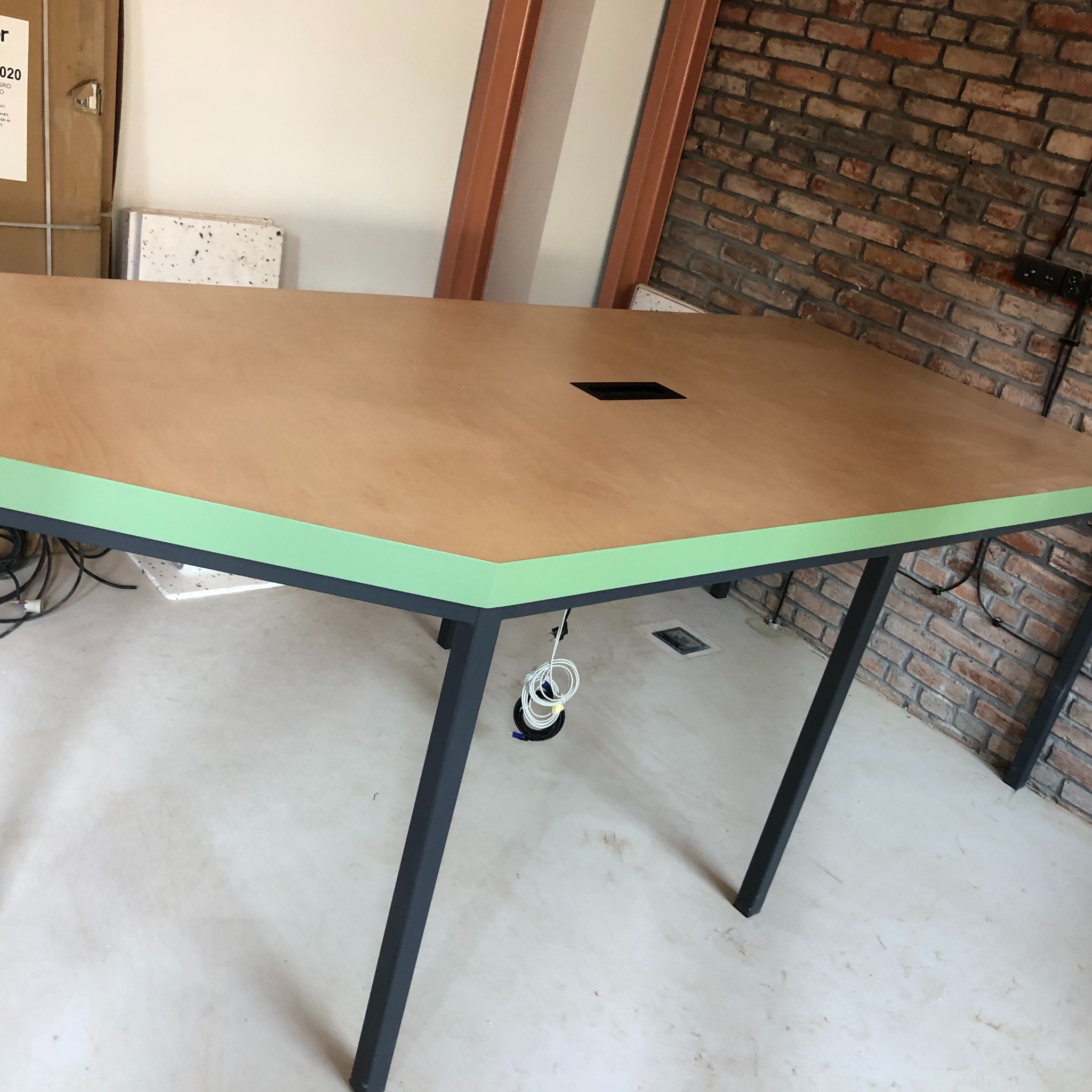 Nietypowy stół konferencyjny o trapezoidalnym blacie z jasnego drewna z zielonym obrzeżem, czarne metalowe nogi, otwór na kable w blacie, widoczna ściana z cegły w tle.