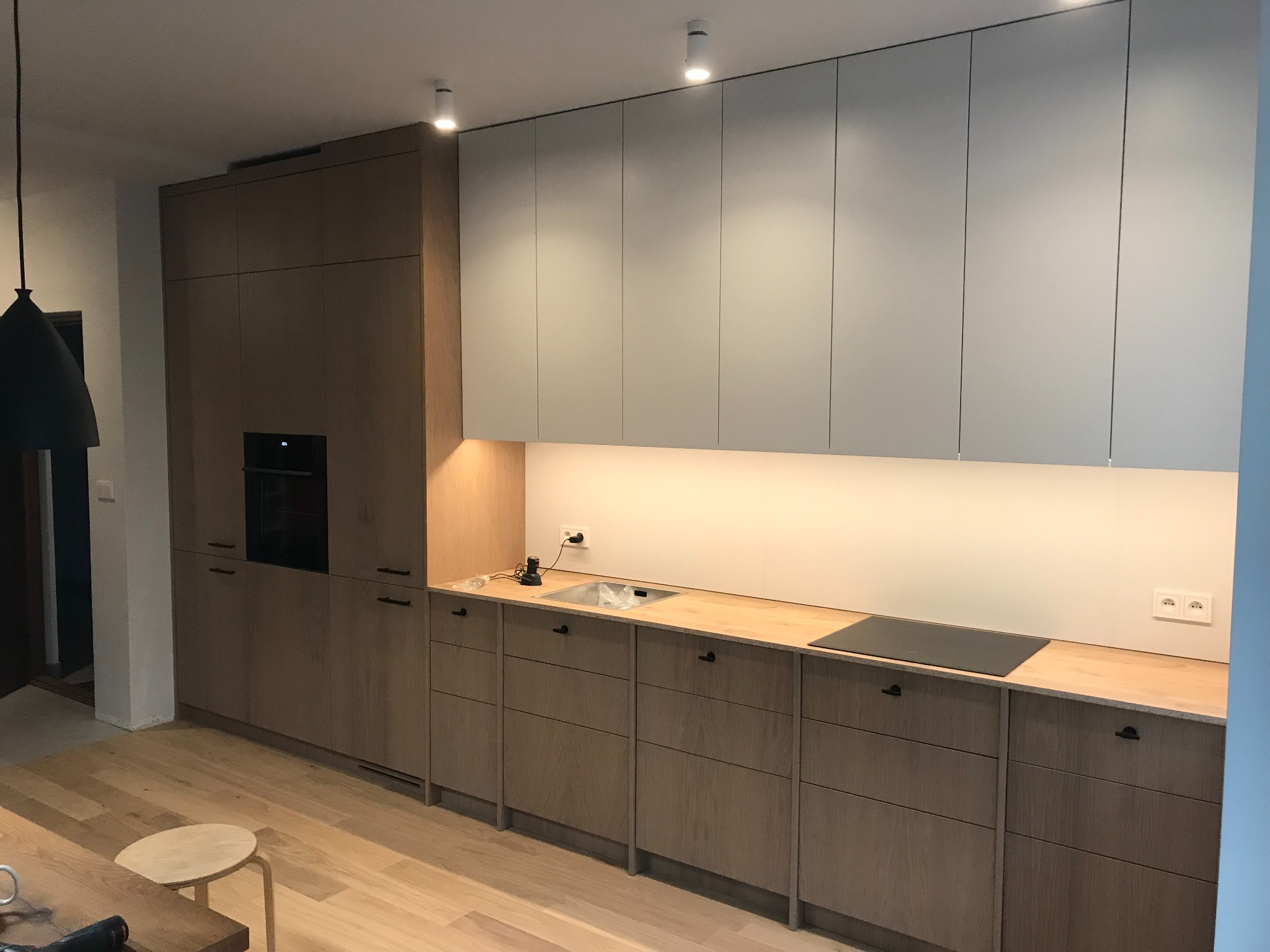 Nowoczesna kuchnia z drewnianymi frontami szafek dolnych i białymi szafkami górnymi, drewniany blat, płyta indukcyjna i oświetlenie podszafkowe LED.