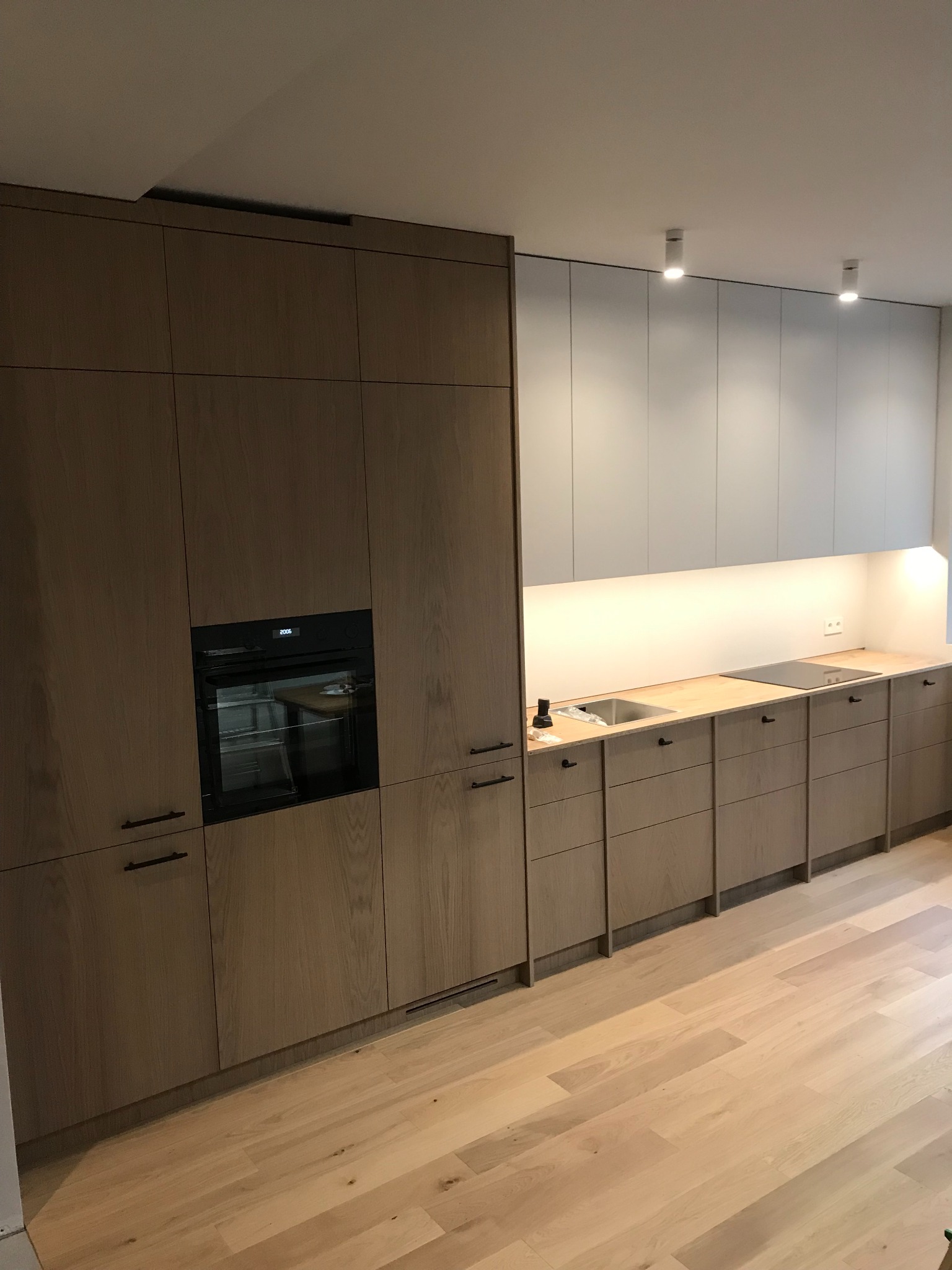 Nowoczesna kuchnia z drewnianymi frontami szafek, czarnymi uchwytami i wbudowanym piekarnikiem, oświetlona listwą LED pod szafkami wiszącymi.