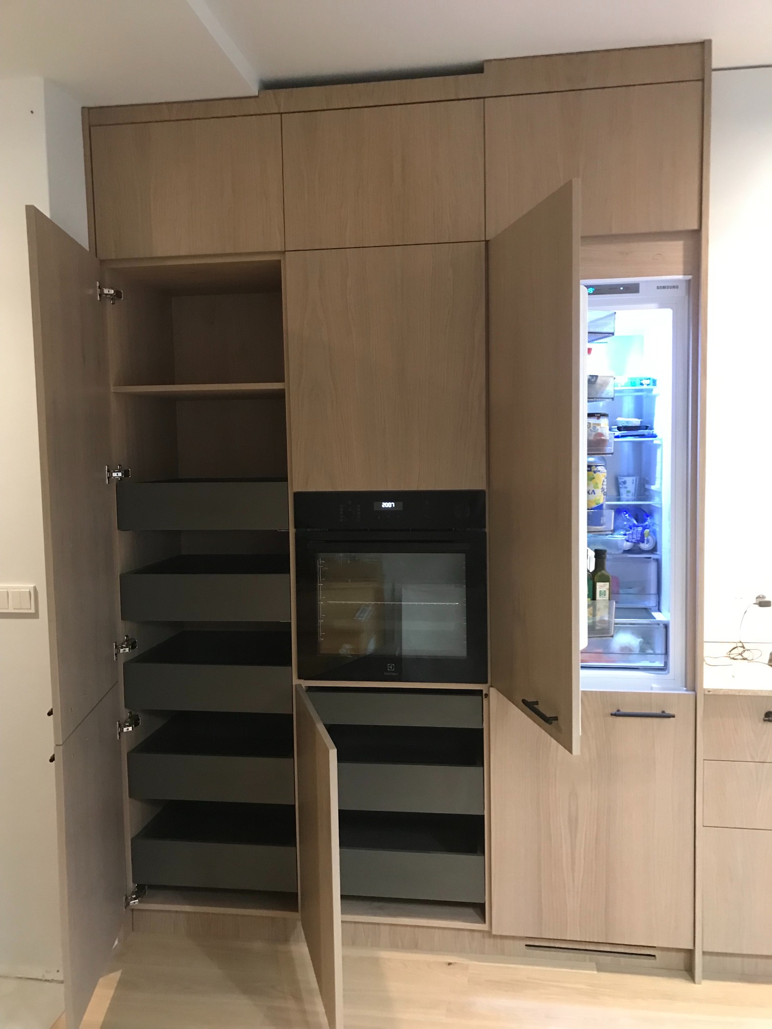 Jasna, drewniana zabudowa kuchenna na wymiar z otwartymi szafkami i szufladami w kolorze grafitowym, piekarnik Electrolux i lodówka Samsung w zabudowie.