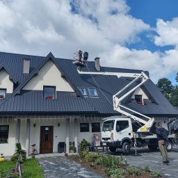 Magdalena WOJTAROWICZ- MAPI - Pracownik na podnośniku koszowym pracuje na dachu domu z blachy trapezowej w Poroninie, widoczny fragment elewacji, okna z kwiatami i zaparkowany samochód firmowy z logo.