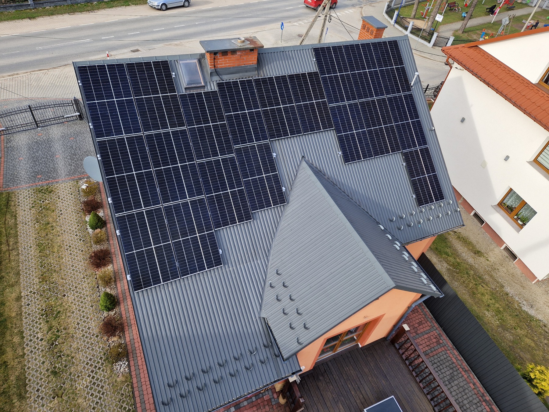 Widok z góry na dom w Niedzicy z panelami słonecznymi na szarym, blaszanym dachu, obok komin z cegły i okno dachowe. W tle ulica, chodnik z kostki i zieleń.