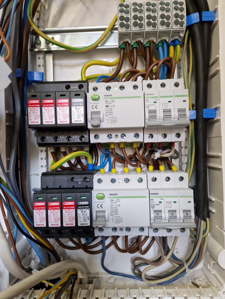 Otwarta skrzynka elektryczna z zainstalowanymi zabezpieczeniami przepięciowymi Phoenix Contact Valvetrab, wyłącznikami nadprądowymi Noark oraz przewodami w różnych kolorach, widoczne oznaczenia L1,...