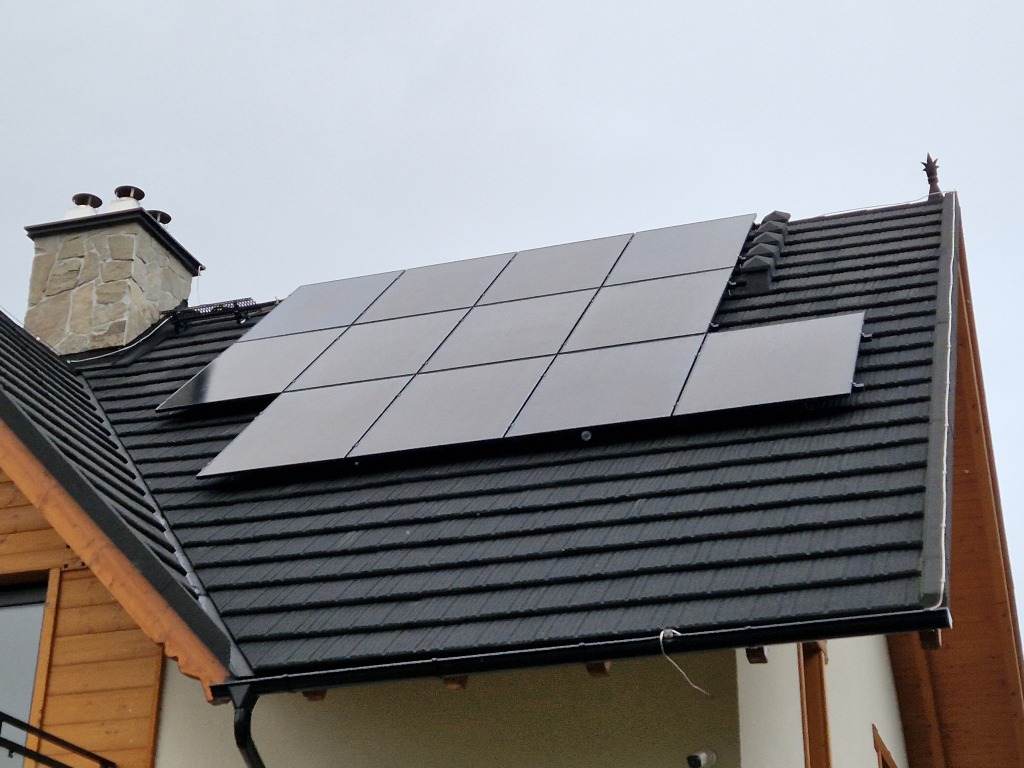 Panele fotowoltaiczne na dachu domu w Szlachtowej, widoczne z boku, z kominem z kamienia w tle.