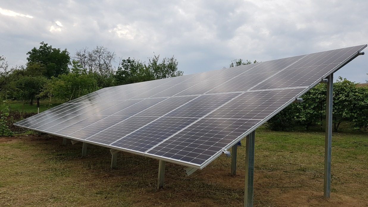 Rząd paneli słonecznych na metalowych słupkach w Olszance, widok z dołu, pochmurne niebo w tle.