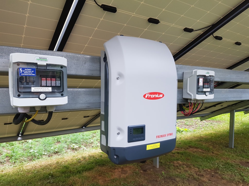 Instalacja fotowoltaiczna: widok od spodu paneli słonecznych na falownik Fronius Symo z wyświetlaczem wskazującym 783, obok skrzynki z bezpiecznikami i okablowaniem, na tle zielonej trawy.