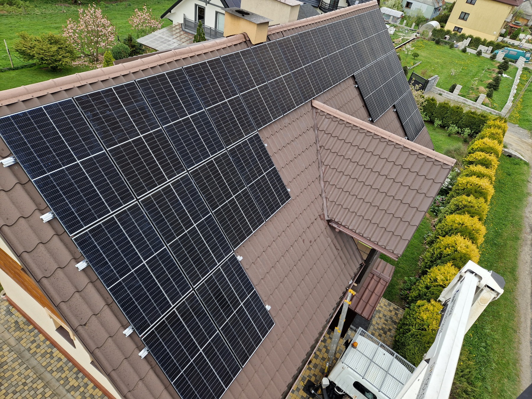 Widok z góry na brązowy dach z panelami słonecznymi; w tle zadbany ogród i zabudowa jednorodzinna, z boku widoczny podnośnik koszowy.
