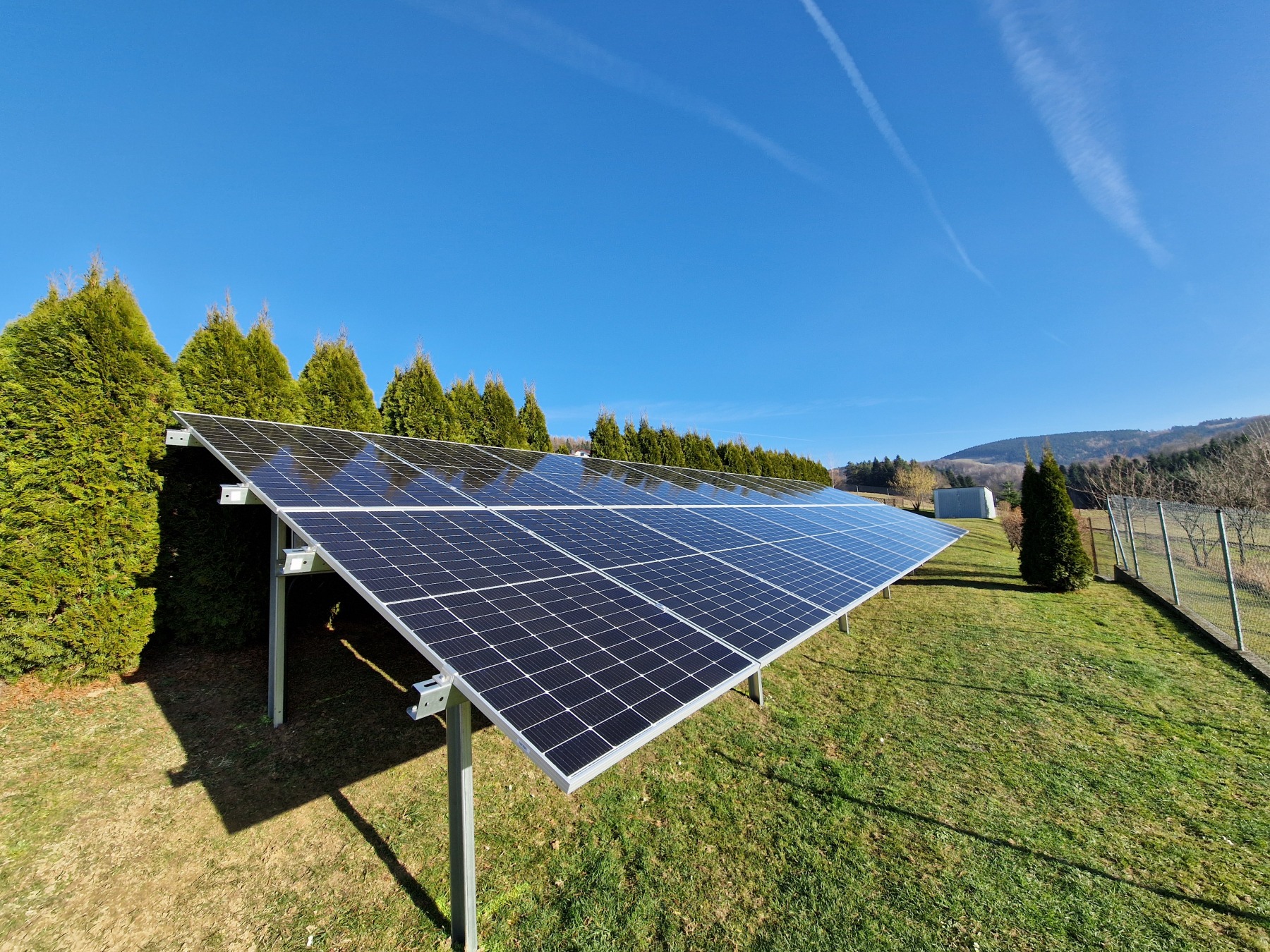 Panel słoneczny na metalowej konstrukcji, z widocznymi śladami kondensacji na powierzchni, otoczony zielonymi tujami i trawnikiem, na tle błękitnego nieba z smugami kondensacyjnymi.