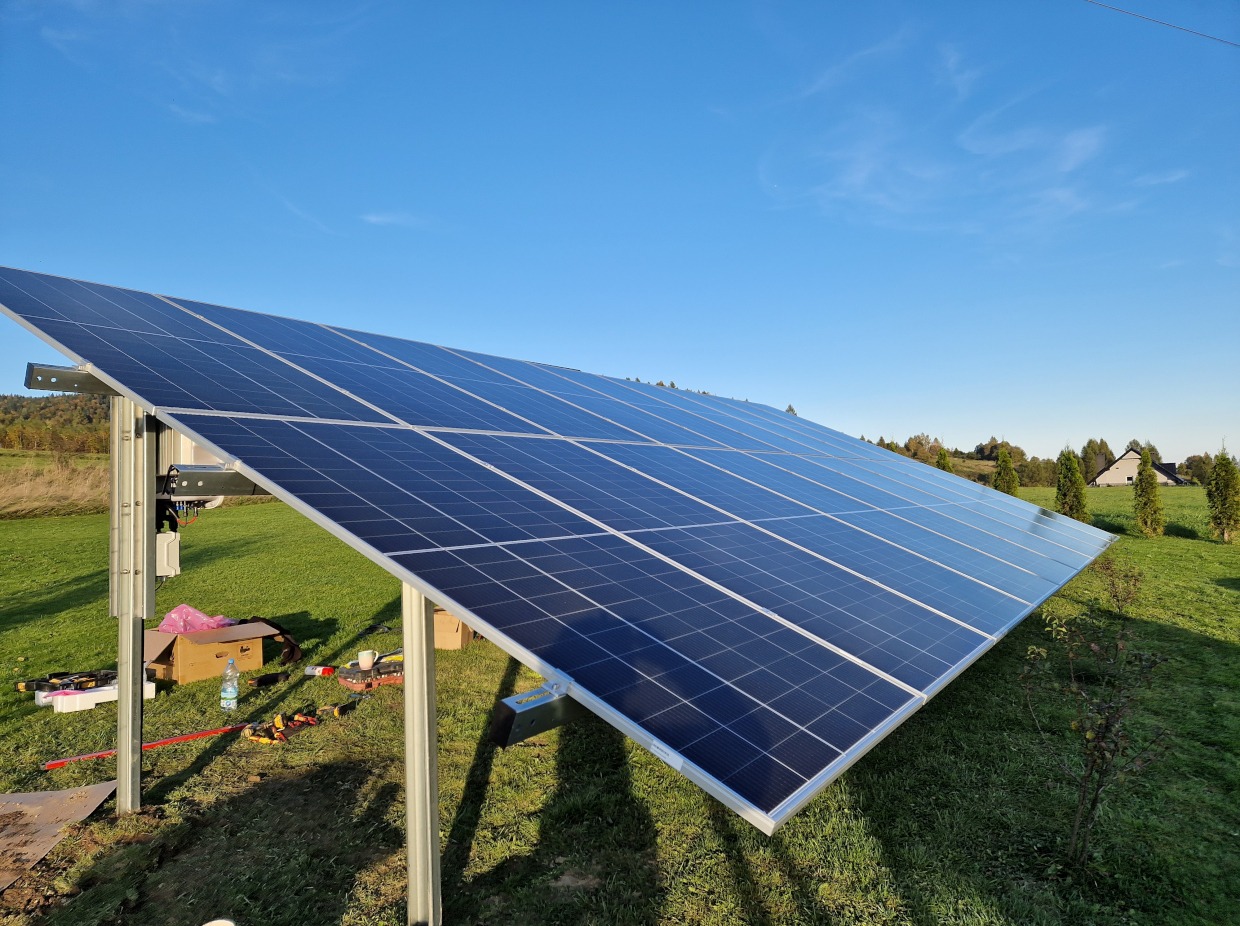 Panele fotowoltaiczne zamontowane na metalowej konstrukcji na trawiastym terenie, widoczne narzędzia i karton, błękitne niebo w tle.