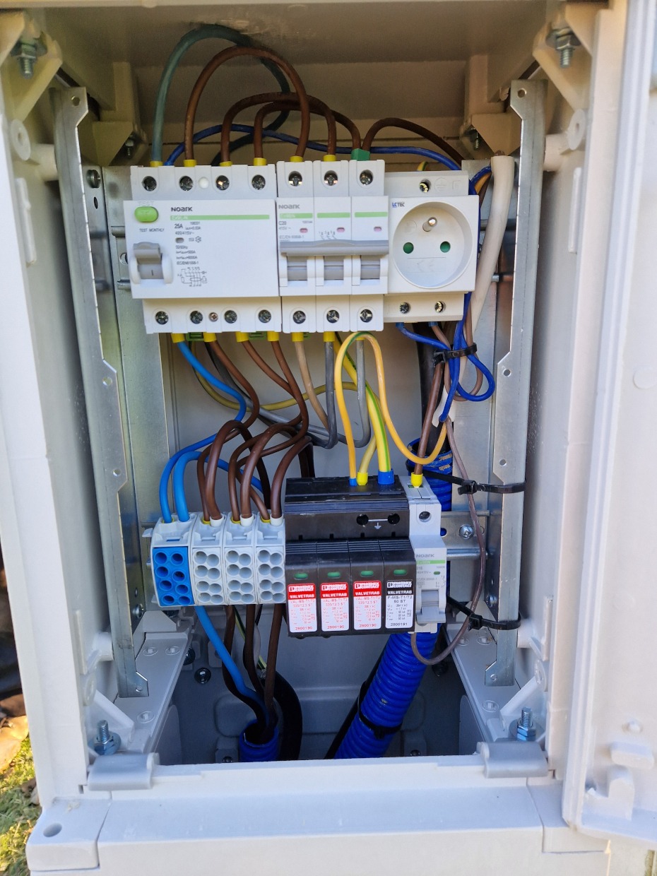 Wnętrze skrzynki elektrycznej z bezpiecznikami, gniazdkiem i przewodami w kolorach brązowym, niebieskim i żółto-zielonym, zabezpieczone niebieską karbowaną rurką.