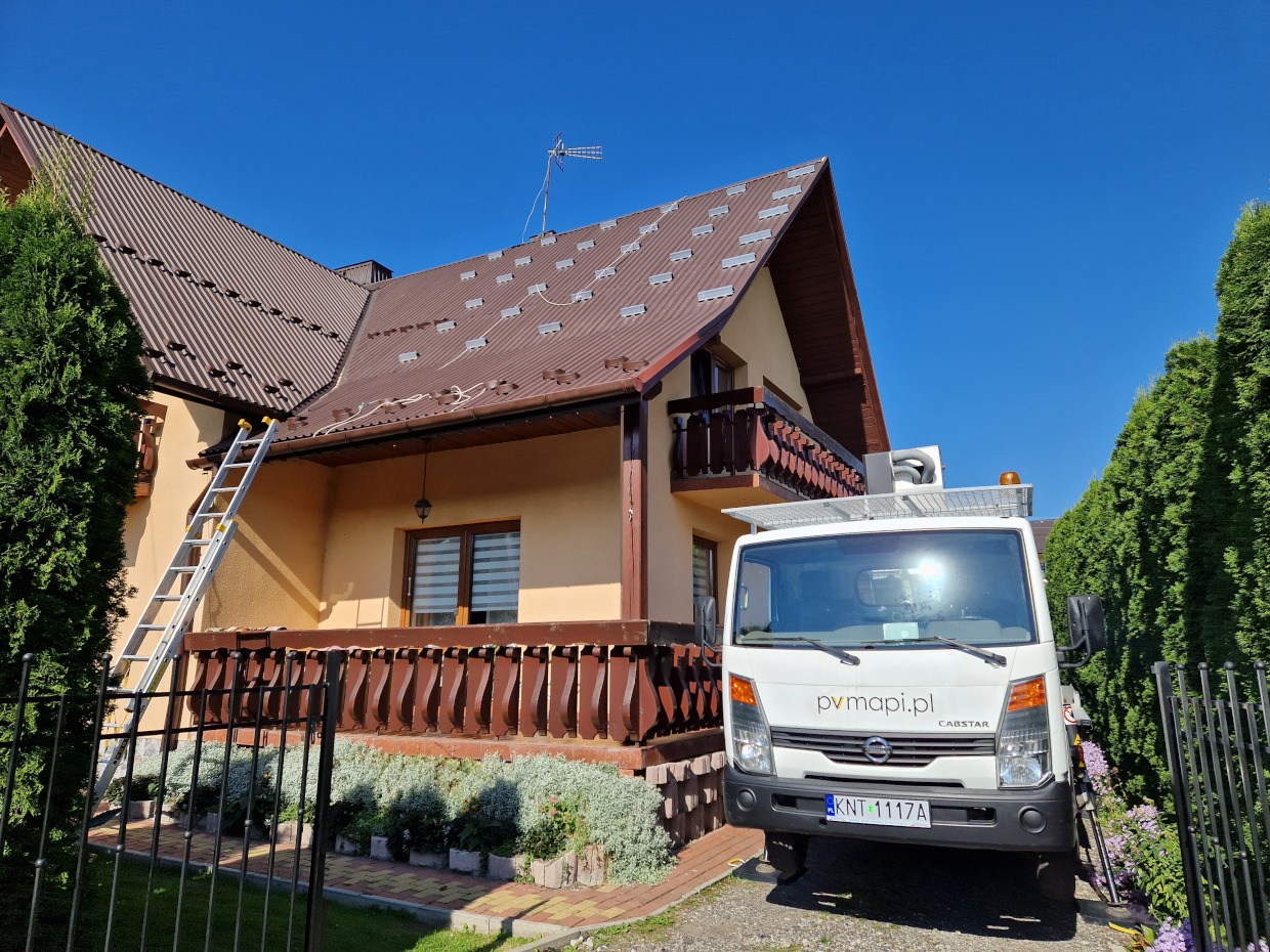 Montaż systemu fotowoltaicznego w Krościenku nad Dunajcem: widok na brązowy dach z zamontowanymi uchwytami pod panele, drabina oparta o ścianę domu, samochód dostawczy z logo firmy na tle zieleni.