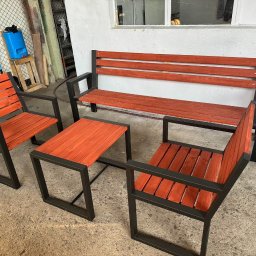 SPAWSTAL VASYL PIDKOPAI - Komplet mebli ogrodowych: ławka, dwa krzesła i stolik z drewnianymi siedziskami i blatami w odcieniach czerwieni, osadzonych na czarnych, metalowych ramach.