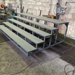 SPAWSTAL VASYL PIDKOPAI - Metalowa konstrukcja schodkowa z drewnianymi siedziskami w warsztacie, widoczne narzędzia i surowy beton na podłodze. Szara kolorystyka i prosta forma.