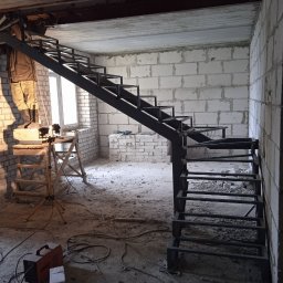 SPAWSTAL VASYL PIDKOPAI - Montaż metalowych schodów w surowym wnętrzu. Widoczny spawacz na podeście, ceglane ściany i sprzęt spawalniczy. Schody o konstrukcji stalowej, jeszcze bez wykończenia.