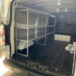 SPAWSTAL VASYL PIDKOPAI - Wnętrze furgonetki z zamontowanym, metalowym regałem na narzędzia i materiały budowlane. Widoczne białe kanistry i worek z zaprawą.