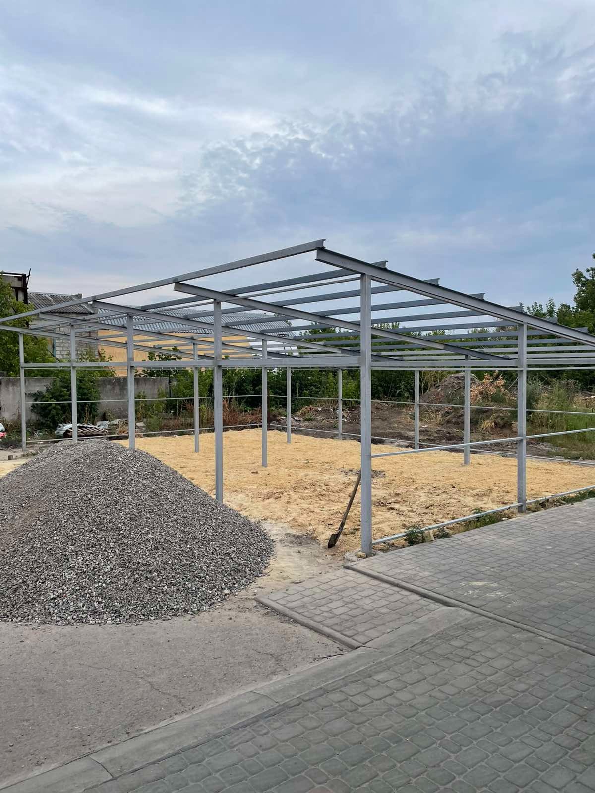 Stalowa konstrukcja hali w trakcie budowy, widoczny szkielet z belek i słupów, obok hałda żwiru i łopata. Teren przygotowany pod wylewkę.