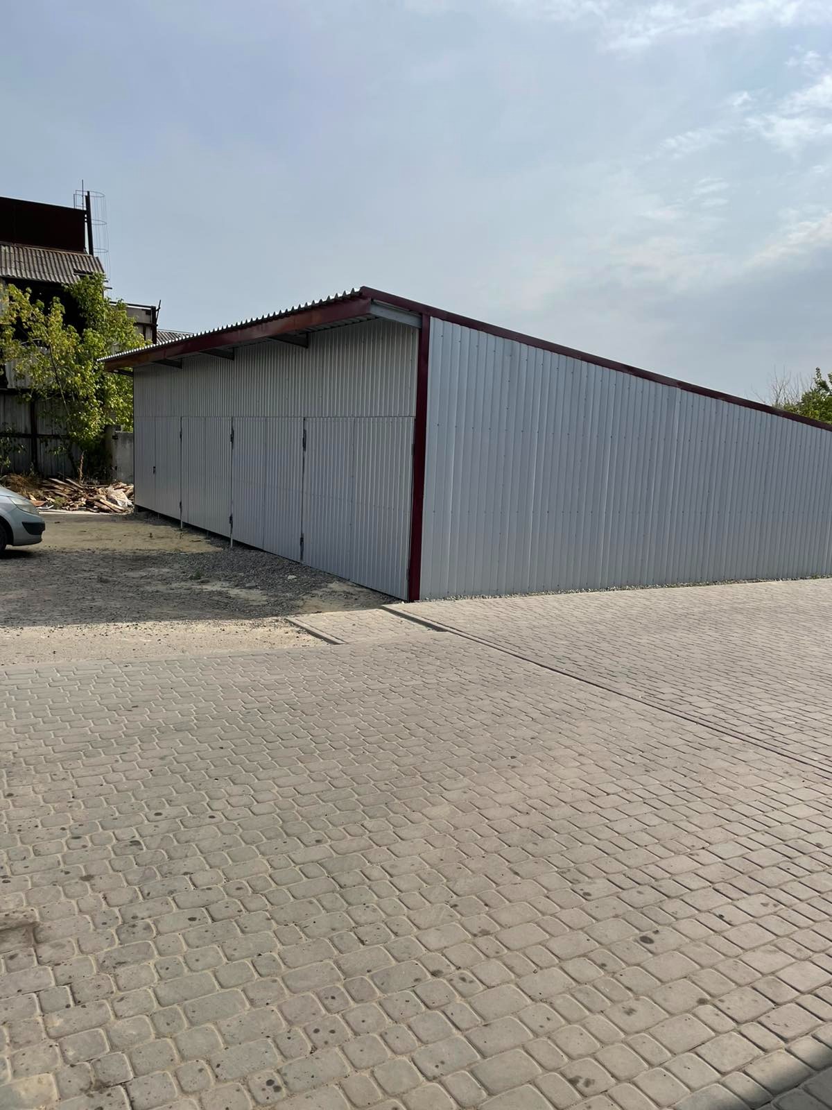 Metalowa hala z czterema bramami garażowymi, wykończona czerwoną ramą, na tle kostki brukowej i zieleni. Widok z zewnątrz, w słoneczny dzień.