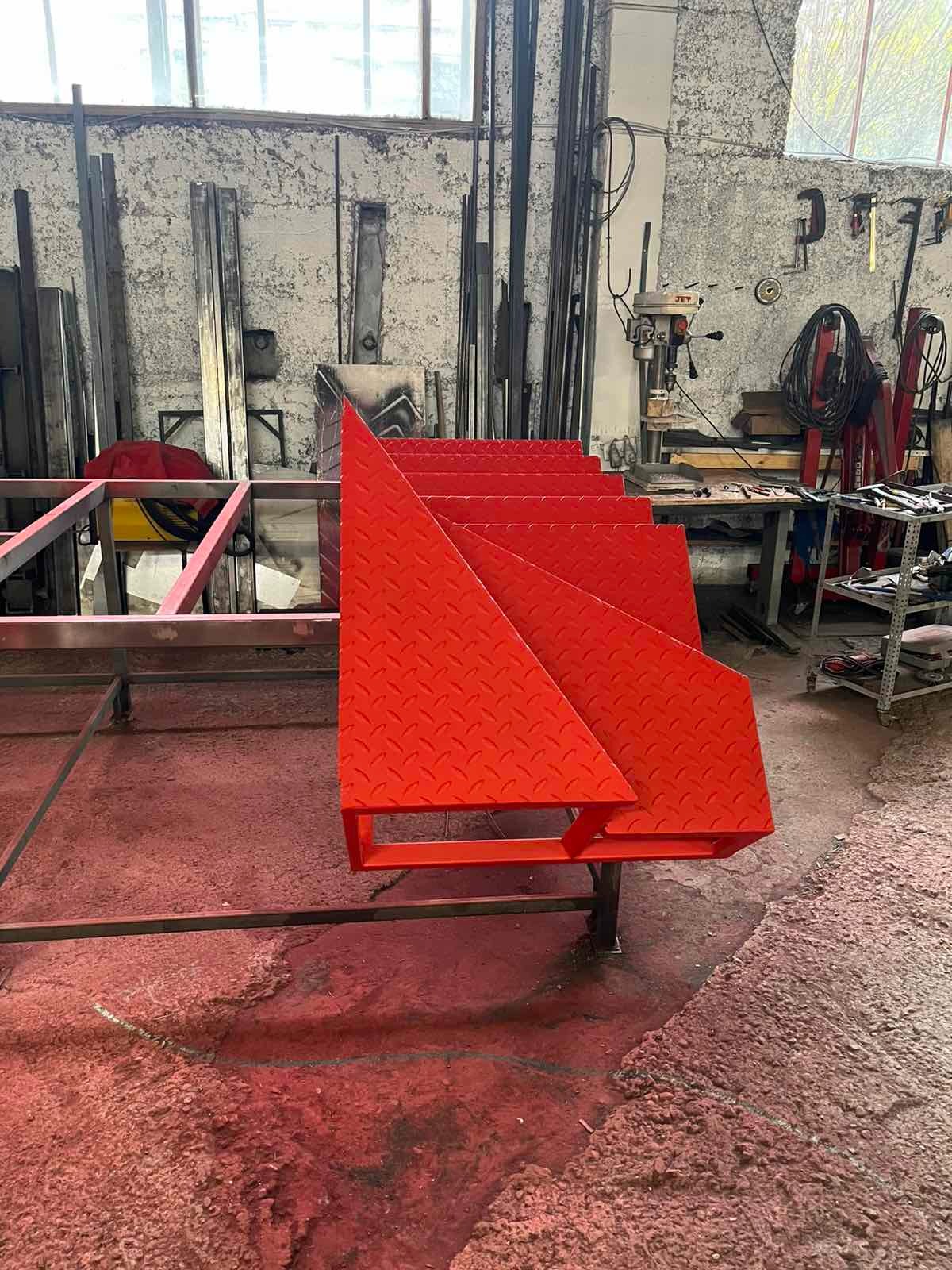Czerwone, metalowe schody z blachy ryflowanej w warsztacie, widoczne narzędzia i elementy konstrukcyjne w tle. Nietypowe ujęcie z dołu, podkreślające geometrię stopni.