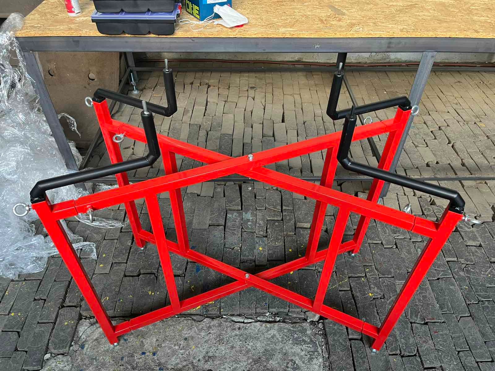 Czerwony, metalowy stojak spawalniczy z czarnymi uchwytami, widoczny na tle podłogi z cegieł i stołu warsztatowego. Konstrukcja spawana, stabilna, z elementami mocującymi.