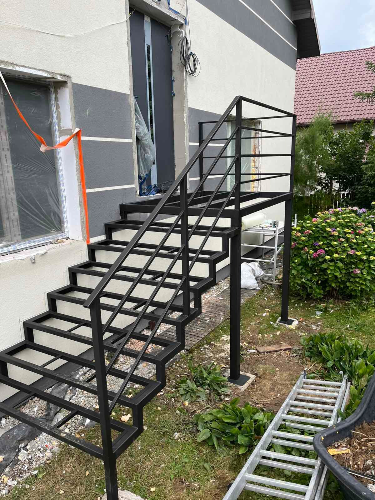 Surowe, czarne, metalowe schody zewnętrzne z poręczą, prowadzące do drzwi budynku. Widoczne elementy montażowe i otoczenie w trakcie prac wykończeniowych.