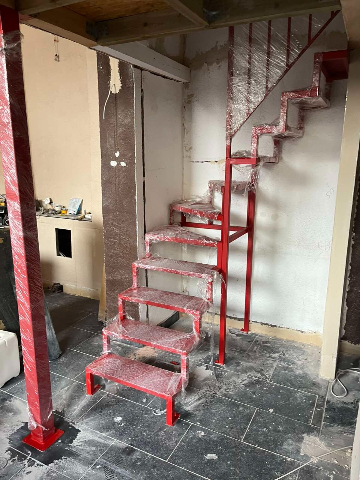 Czerwone, metalowe schody modułowe w folii ochronnej, ustawione w surowym wnętrzu. Widoczny fragment słupa nośnego i fragment podłogi z ciemnych płytek.
