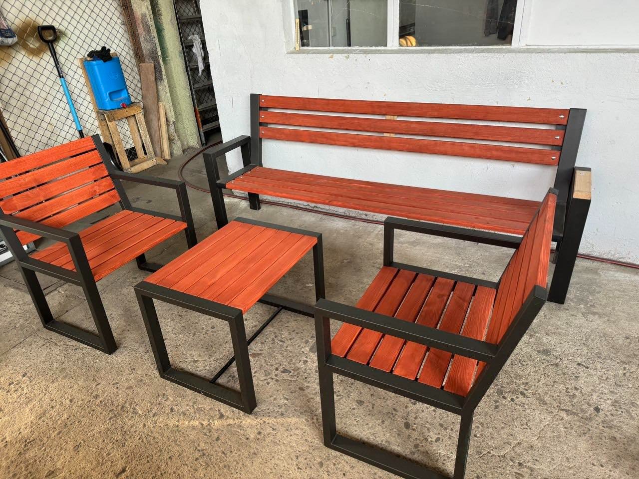 Komplet mebli ogrodowych: ławka, dwa krzesła i stolik z drewnianymi siedziskami i blatami w odcieniach czerwieni, osadzonych na czarnych, metalowych ramach.