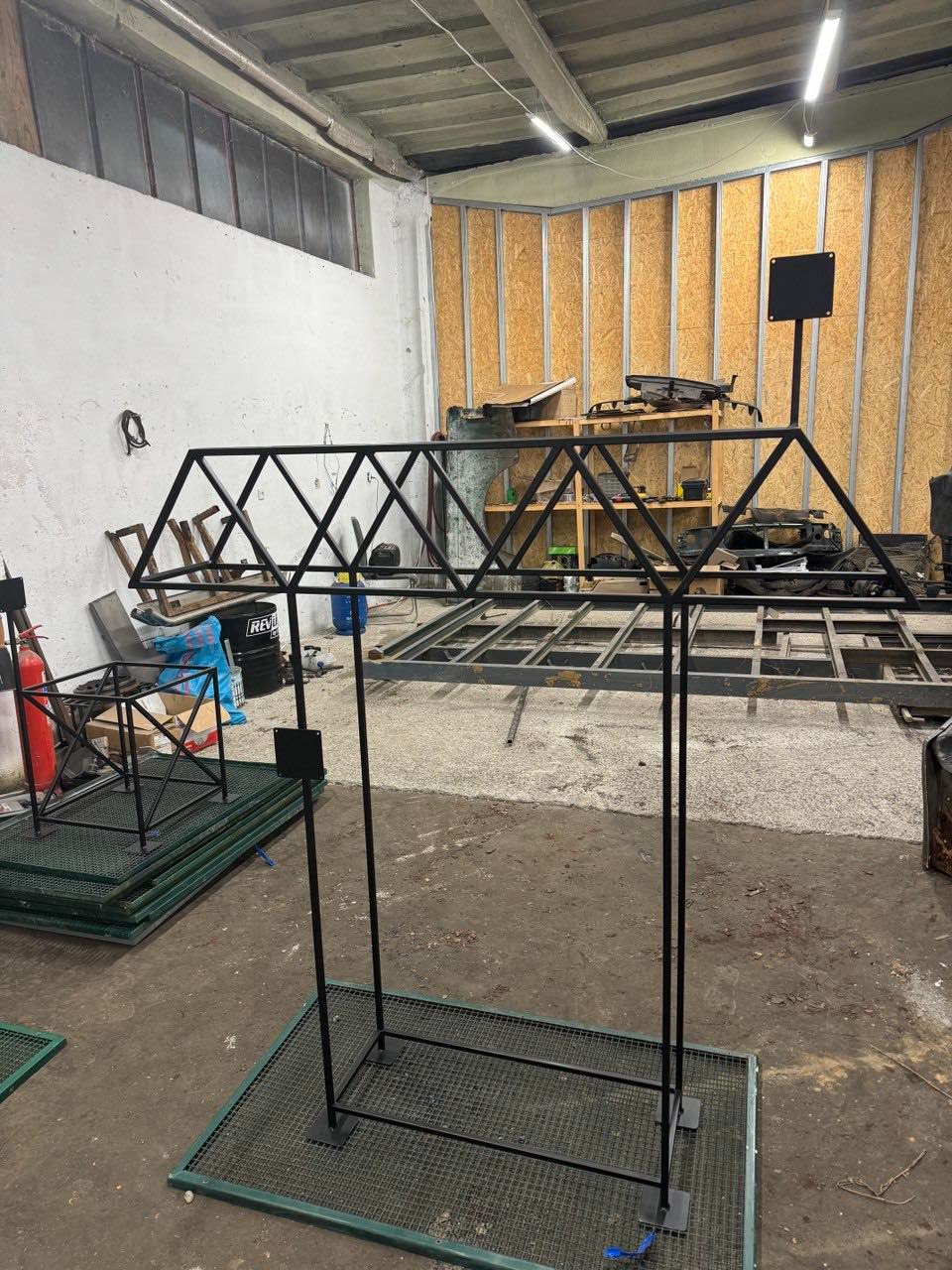 Konstrukcja metalowa w warsztacie: czarna rama z trójkątnymi wzmocnieniami, spawana na metalowej siatce, surowy beton i drewniane panele w tle.