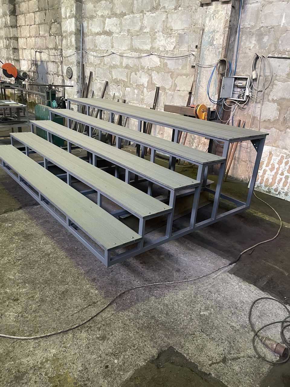 Metalowa konstrukcja schodkowa z drewnianymi siedziskami w warsztacie, widoczne narzędzia i surowy beton na podłodze. Szara kolorystyka i prosta forma.