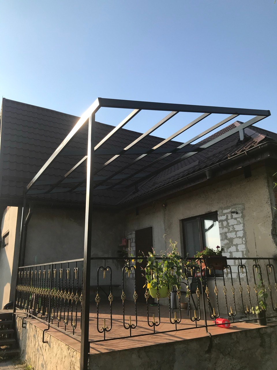 Metalowa konstrukcja z poręczą na tarasie domu w słoneczny dzień. Czarna, ażurowa balustrada z dekoracyjnymi elementami. Rośliny w doniczkach dodają uroku.