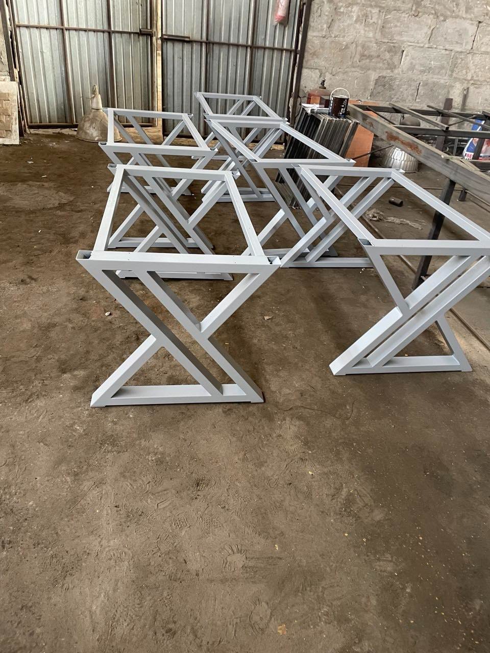 Metalowe, pomalowane na szaro, spawane podstawy stołów z geometrycznym wzorem X, ustawione w warsztacie. Widoczne ślady pracy na podłodze.
